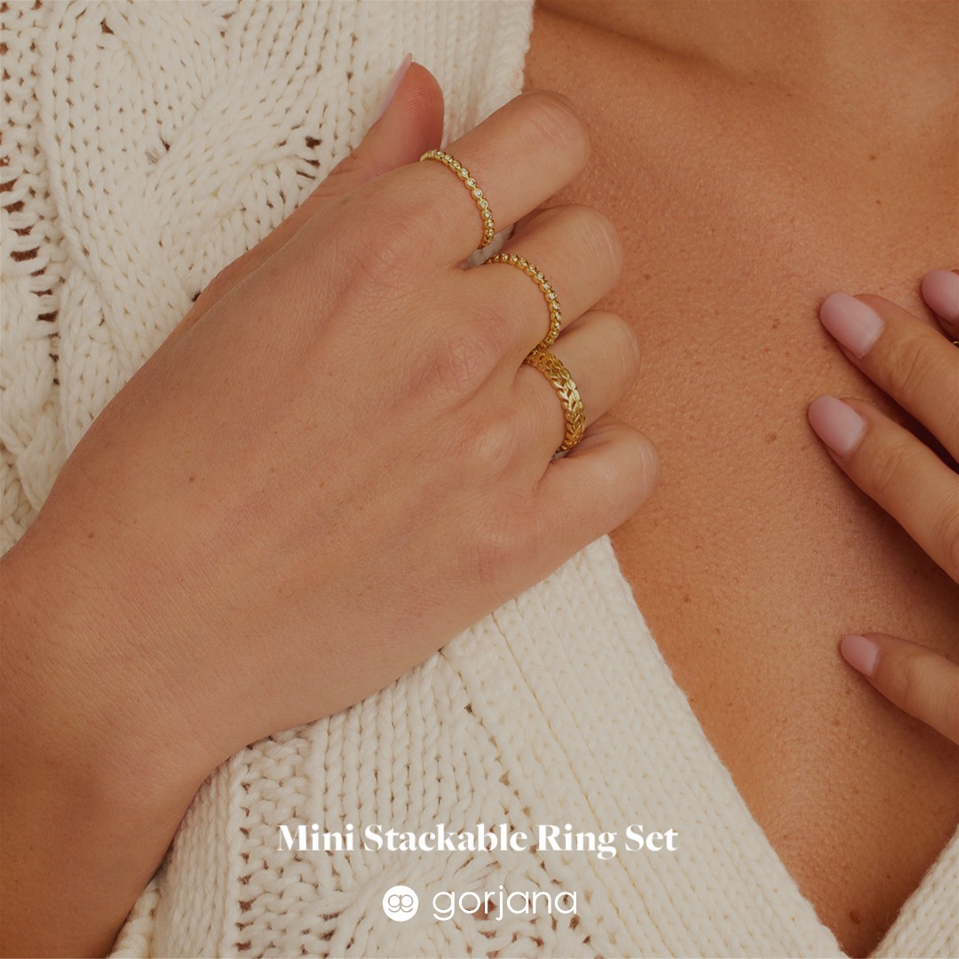 A close-up shot focuses on a person's hand adorned with three delicate gold rings, stacked on one finger. The hand rests against a cream-colored, textured knit sweater. Overlayed on the bottom of the image is the text "Mini Stackable Ring Set" followed by the Gorjana logo. The overall aesthetic is elegant and minimalist, highlighting the jewelry.