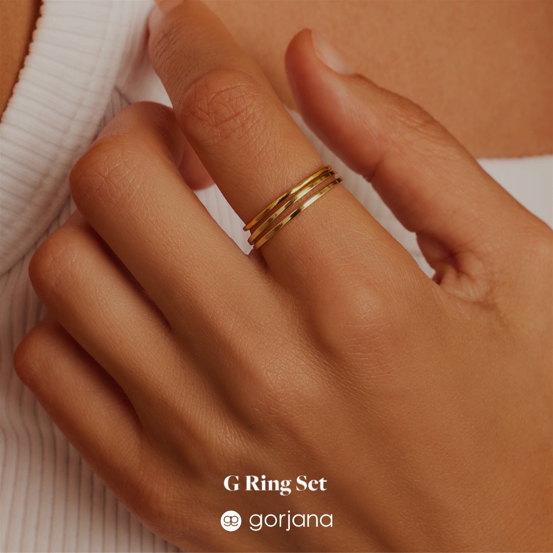 A close-up shot focuses on a hand adorned with three delicate gold rings stacked on one finger. The background is softly blurred, highlighting the jewelry. White text overlaid on the image reads "G Ring Set" and "gorjana" with a small logo. The overall aesthetic is minimalist and elegant, emphasizing the product's design and brand.