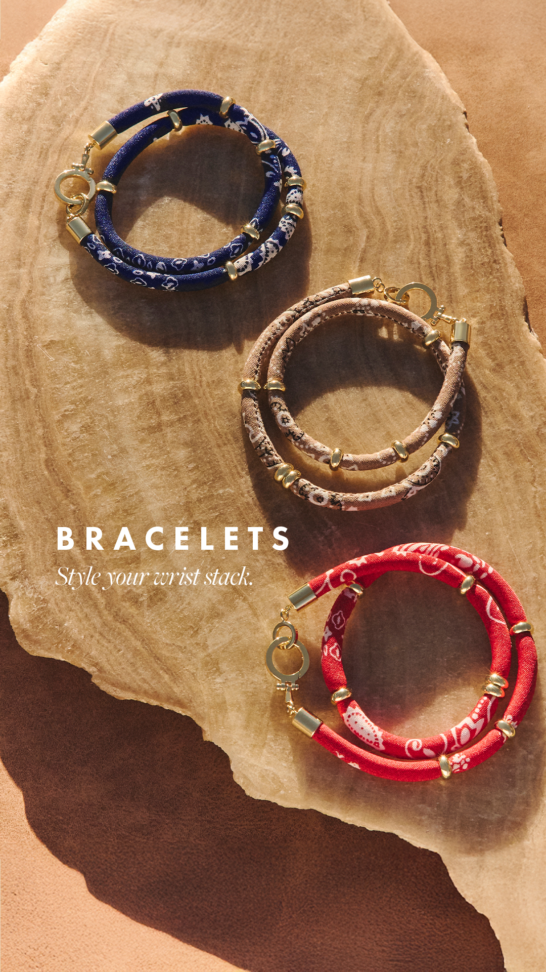 Three fabric bracelets with gold hardware are displayed on a textured, light brown surface. The bracelets are navy blue with a white pattern, tan with a snake-print pattern, and red with a white bandana-style pattern. The text "BRACELETS" is displayed in large white sans-serif font, with the tagline "Style your wrist stack." in a smaller, italicized font below it.