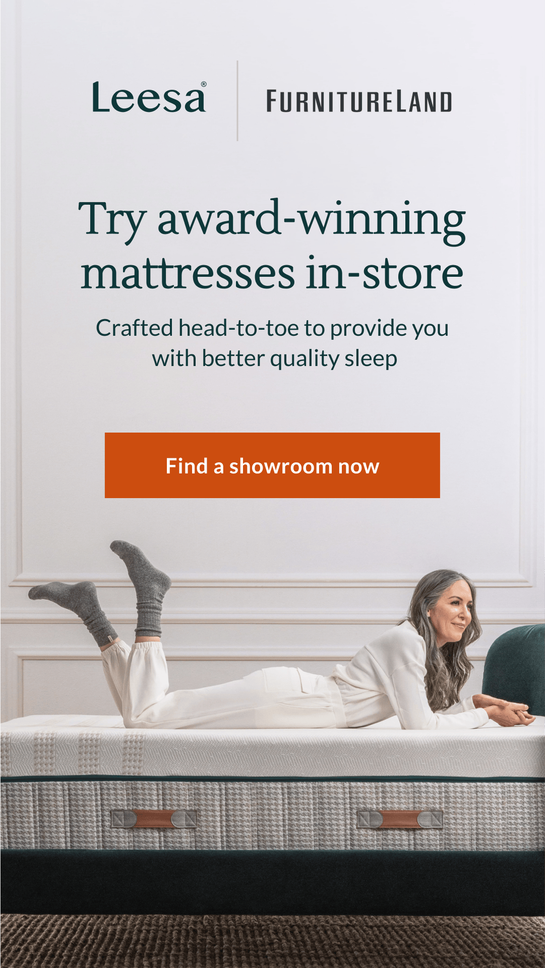 A woman with gray hair is lying on a Leesa mattress, wearing white clothing and gray socks. She is looking to the side with a slight smile. The text "Leesa" and "FURNITURELAND" are at the top of the image. Below that, the headline reads "Try award-winning mattresses in-store" with the sub-headline "Crafted head-to-toe to provide you with better quality sleep." An orange button with the text "Find a showroom now" is visible.