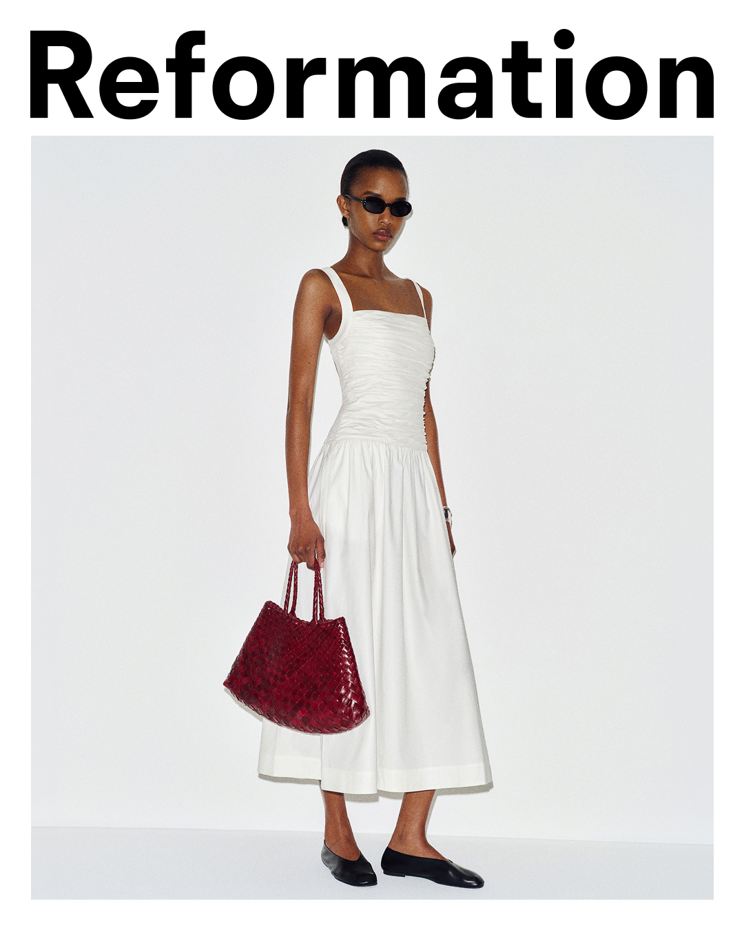 A model stands against a white background wearing a white ruched midi dress and black ballet flats. She holds a large, woven red handbag. The word "Reformation" is displayed in large black text at the top of the image. The text "Not a mini, not yet a maxi. Shop the Nesta Midi Dress." is overlaid on the image, describing the dress.