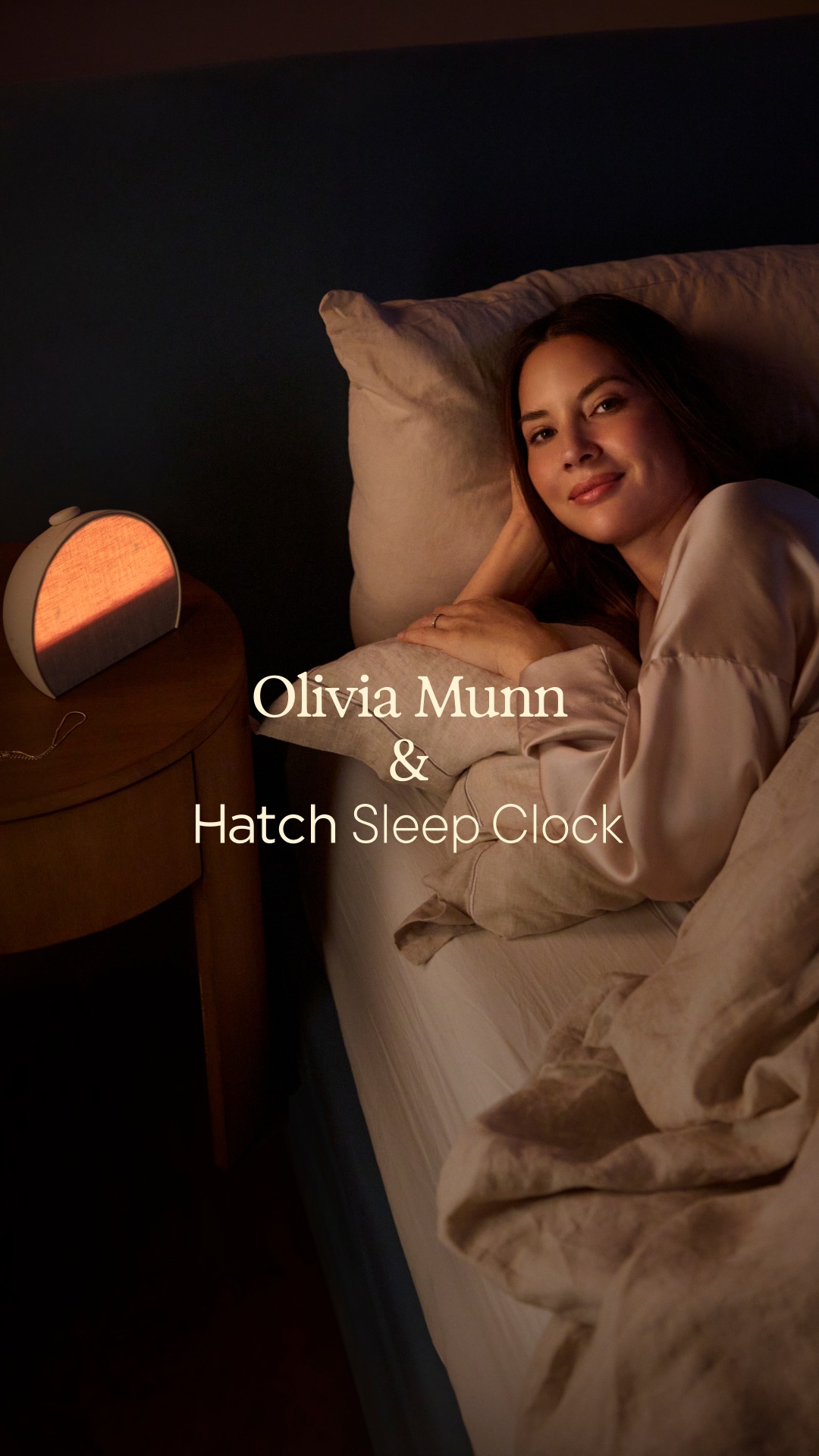 The image features actress Olivia Munn lying in bed, looking relaxed and smiling at the camera. Beside the bed, on a nightstand, is a Hatch Sleep Clock emitting a warm, orange glow. Text overlaid on the image reads "Olivia Munn & Hatch Sleep Clock", highlighting the collaboration. The overall scene evokes a sense of calm and restful sleep, suggesting the product contributes to a peaceful night.