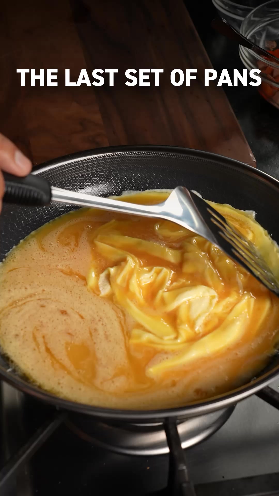 A close-up shot shows eggs being scrambled in a HexClad pan. White text overlaid on the image reads "THE LAST SET OF PANS". The pan has a honeycomb pattern on its surface, and a metal spatula is used to stir the eggs. The scene is focused on the cooking process within the pan.