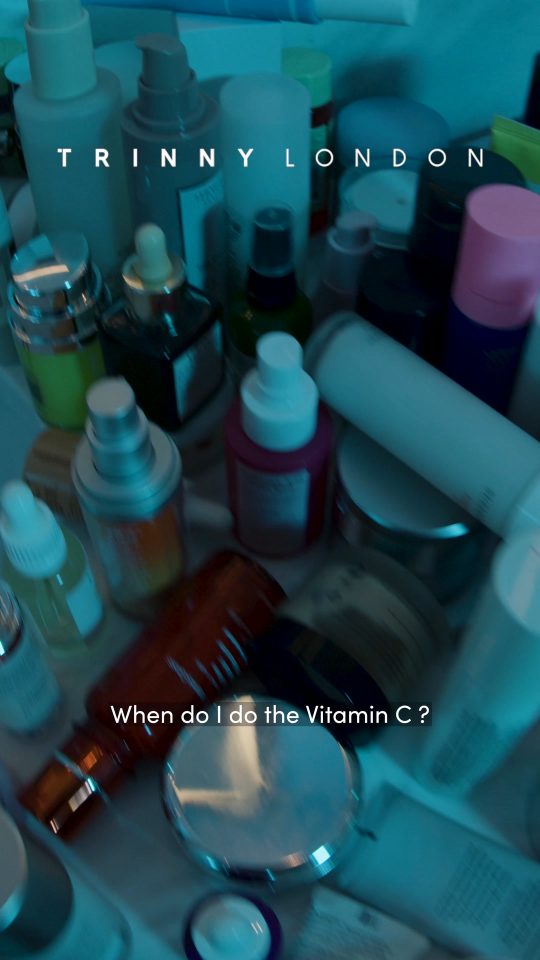 A close-up, slightly blurry shot of numerous skincare bottles and jars arranged together. The brand name "TRINNY LONDON" is visible in white text across the top of the image. White text overlaid on the bottom of the image asks, "When do I do the Vitamin C ?" The overall lighting is dim and has a blueish tint.