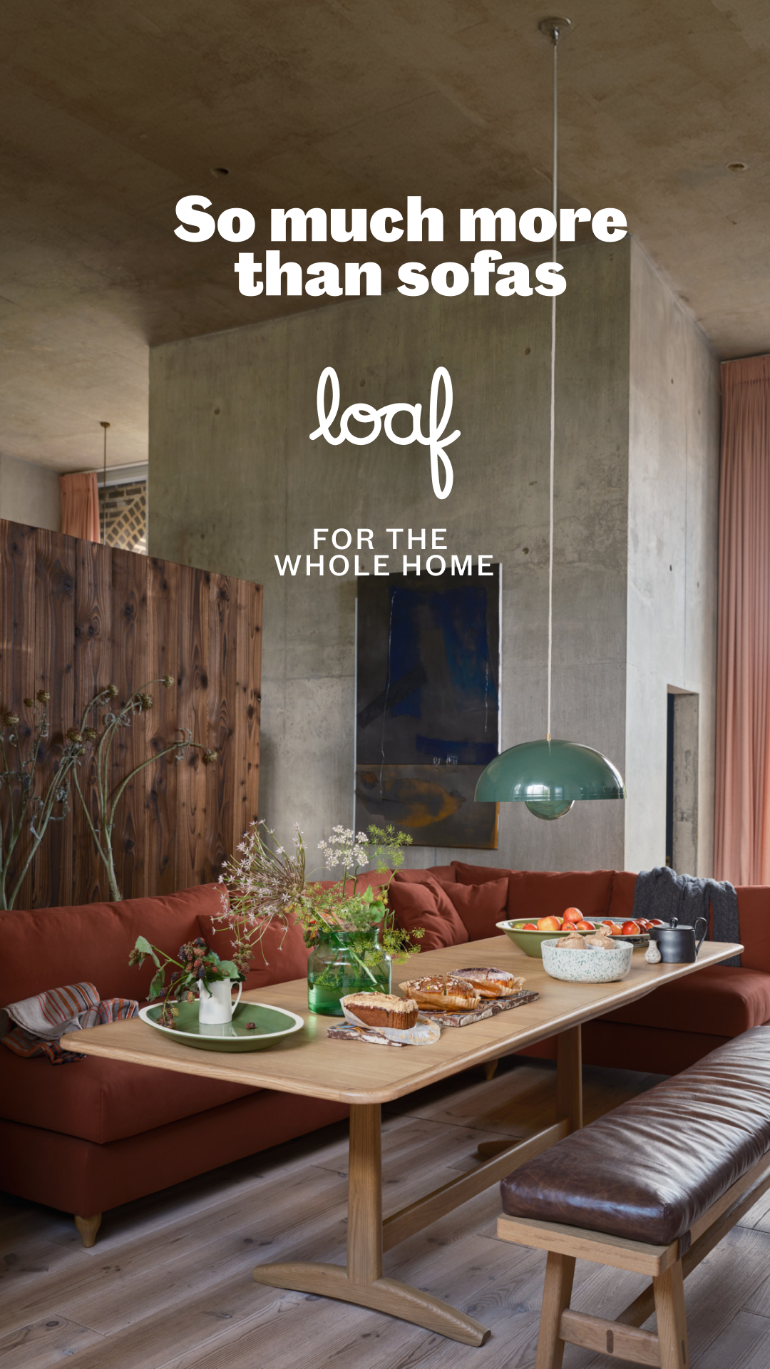 A wide shot of a modern, minimalist living and dining area. A large, rust-colored sectional sofa is arranged around a wooden dining table. On the table, there are pastries, fruit in a bowl, and decorative items. The text "So much more than sofas" is overlaid in large white sans-serif font at the top of the image. Below that, the "loaf" logo is displayed in white script, followed by "FOR THE WHOLE HOME" in smaller white sans-serif text.