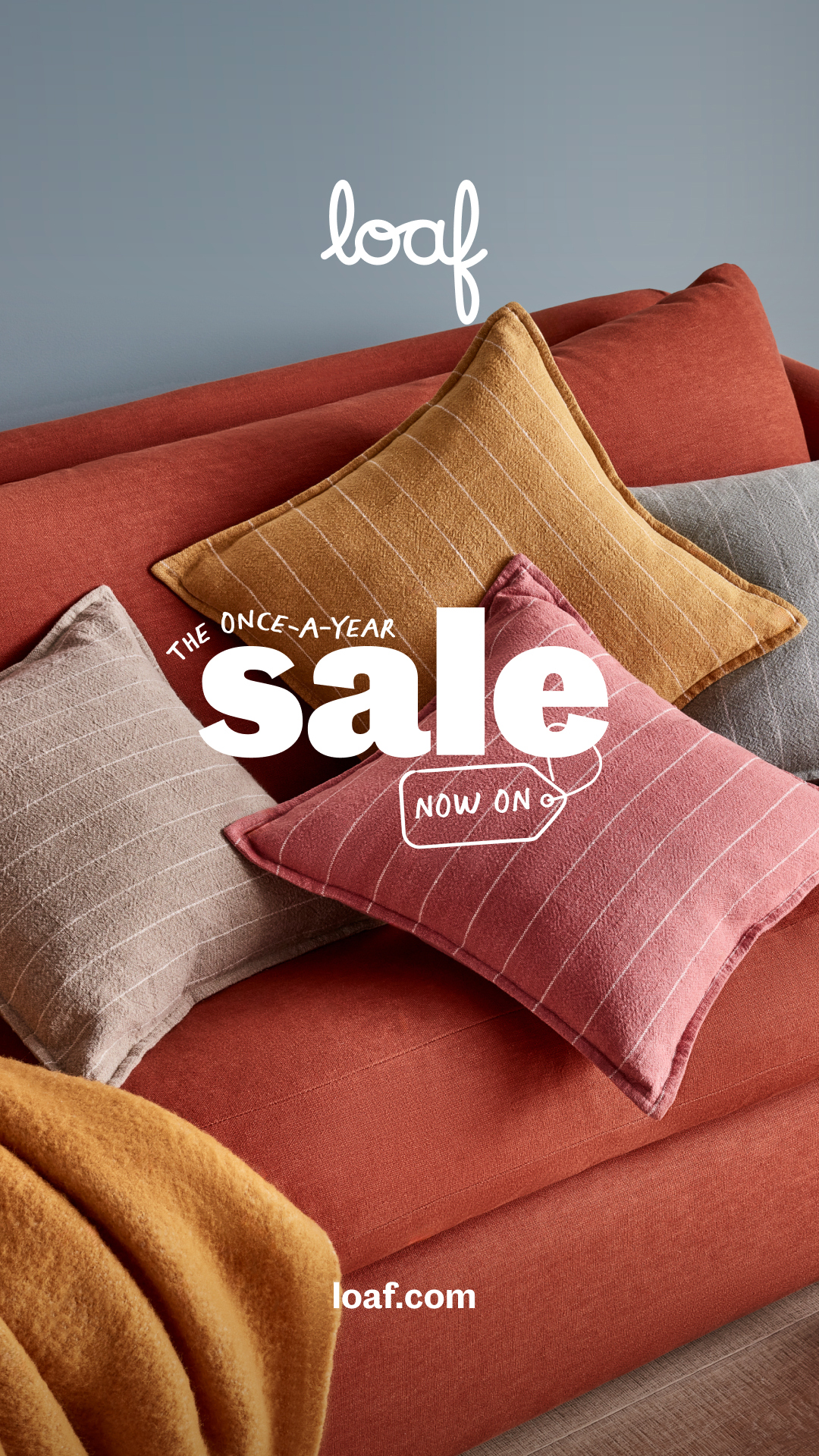 A close-up shot of a sofa with several decorative pillows. The pillows are in shades of rust, mustard yellow, and grey, with subtle striped patterns. White text overlays the image, announcing "THE ONCE-A-YEAR SALE" in large, bold letters, with "NOW ON" inside a tag graphic. The brand logo "loaf" is visible in white script at the top, and "loaf.com" is at the bottom.