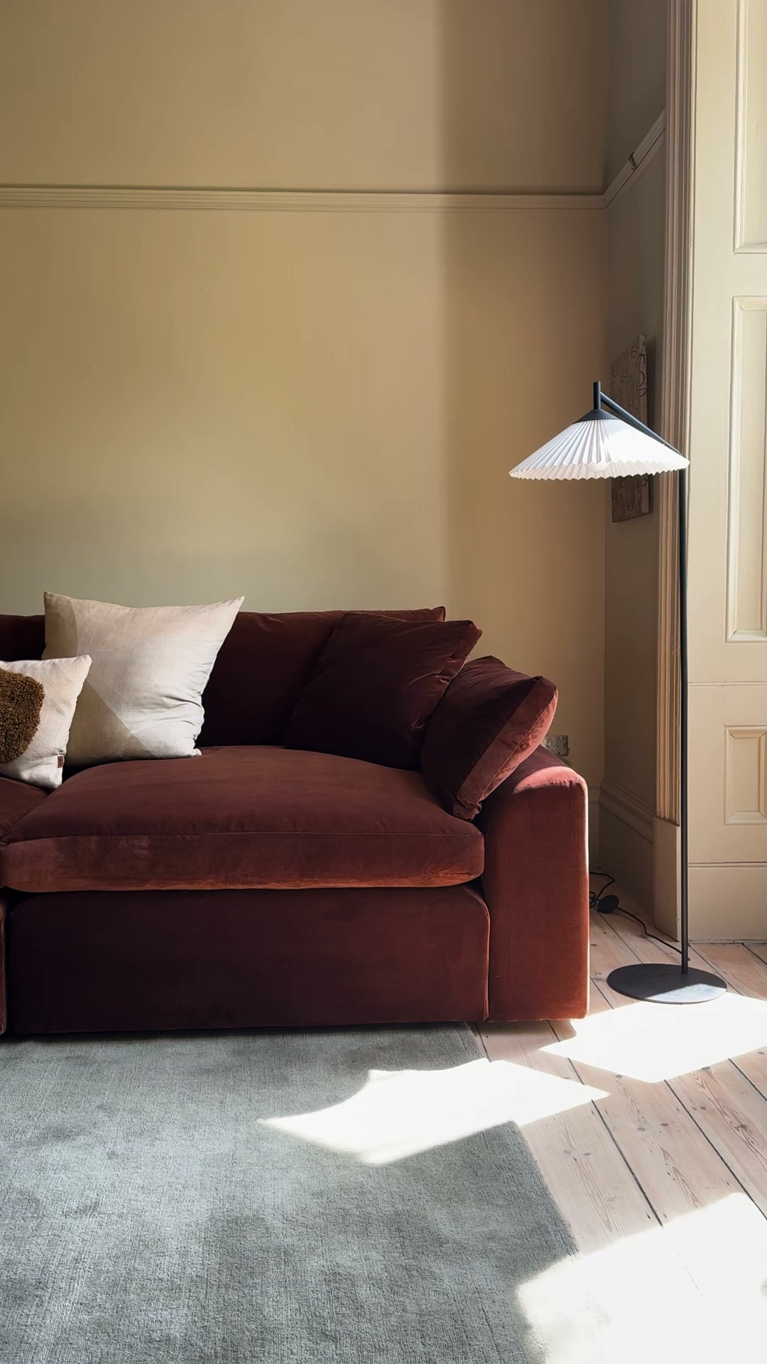 A plush, deep red velvet sofa with several large cushions is shown in a sunlit room. The sofa is placed on a grey rug in front of a pale yellow wall with decorative molding. A tall, modern floor lamp with a pleated white shade stands to the right of the sofa. Sunlight streams in from the right, casting geometric patterns on the wooden floor and the rug. The text "Meet Cuddlemuffin." is overlaid on the image, followed by descriptive text about the sofa's comfort and size.