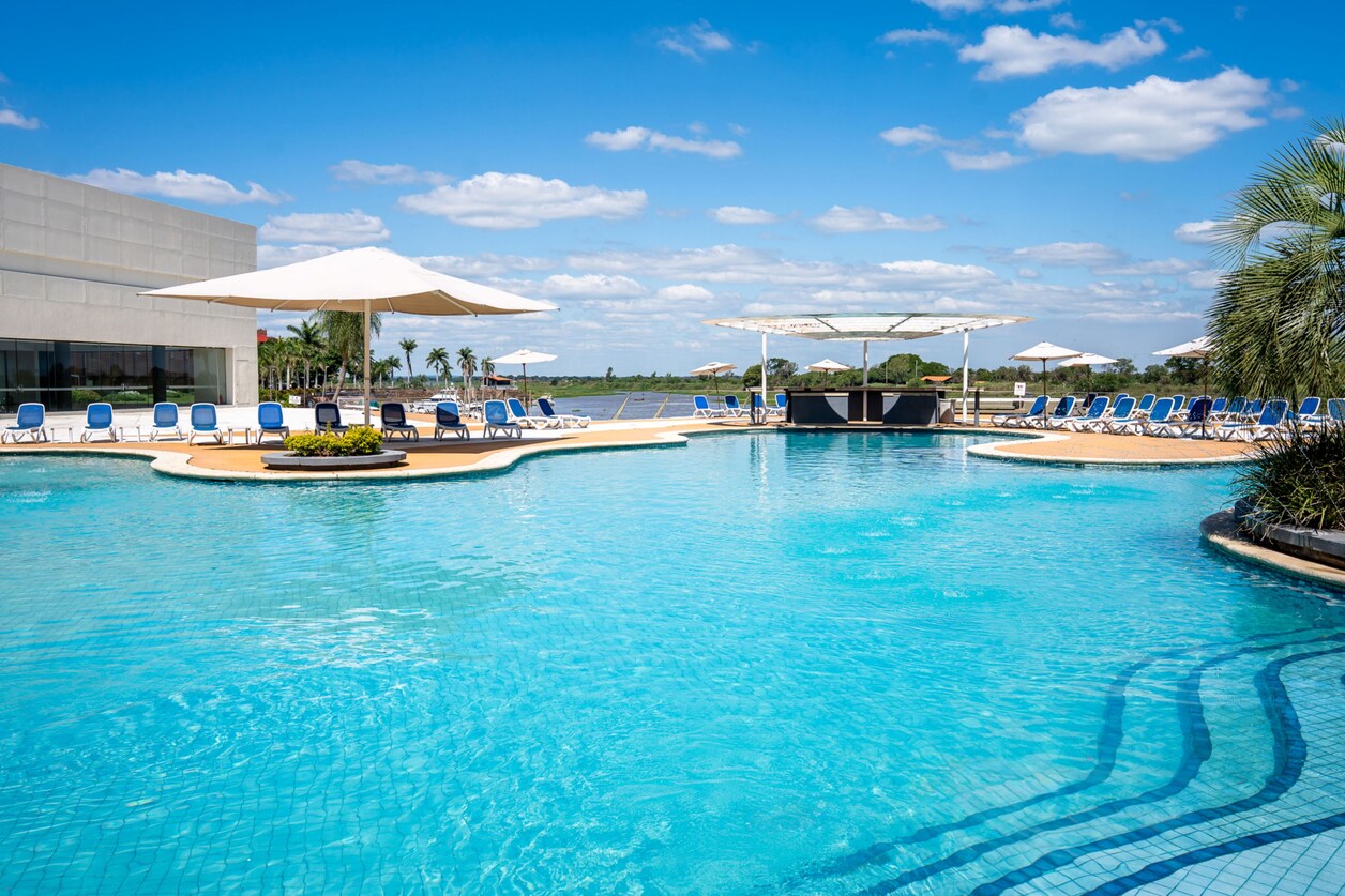 A wide shot of a resort swimming pool on a sunny day. The pool is bright blue and has steps leading into it on the right side. Around the pool are many lounge chairs, some with umbrellas. In the background, there is a bar area and a body of water. Palm trees and a building are visible on the left side. The text overlay reads: "Discover every corner of your favorite destinations with an urban getaway at Yacht & Golf Club Paraguayo, a Tribute Portfolio Resort."