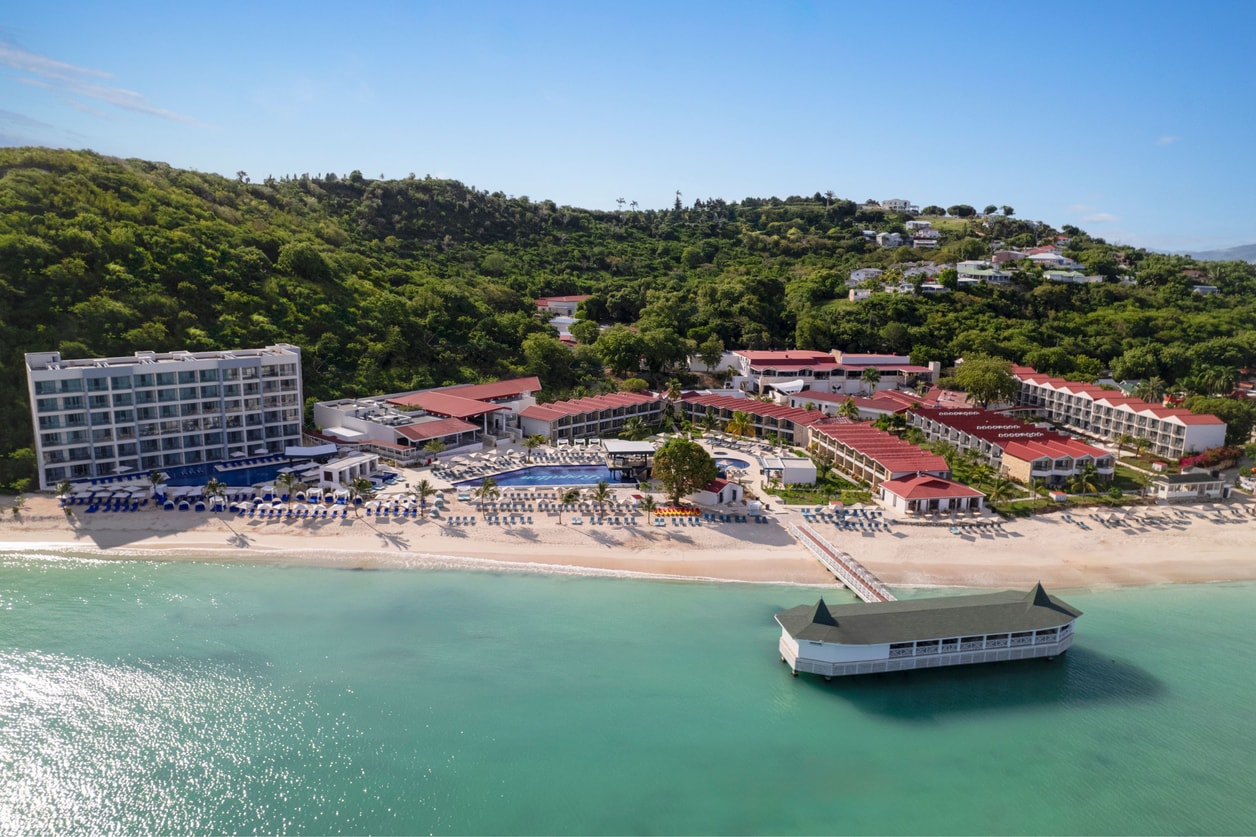 An aerial drone shot showcases a sprawling, luxurious beachfront resort complex under a clear blue sky. The resort features multiple buildings with red roofs and white facades, a large swimming pool, numerous lounge chairs on a sandy beach, and a long pier extending into the turquoise ocean. The text overlay promotes booking a memorable vacation at the adults-only, all-inclusive Royalton CHIC Antigua resort with exclusive member rates.