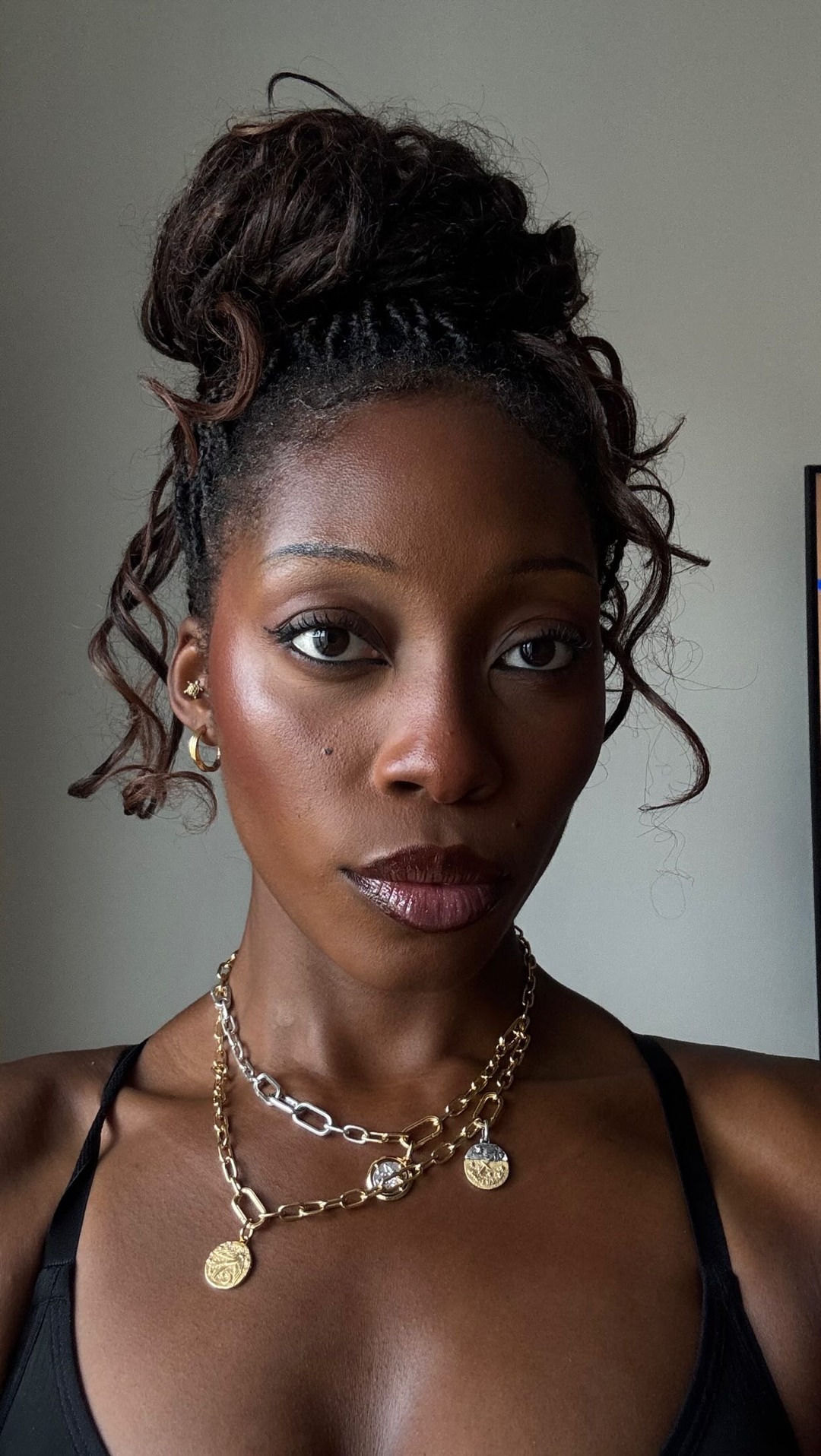 A close-up, head-and-shoulders shot of a Black woman with her hair styled in a messy bun with some curls framing her face. She is wearing a black tank top and layered gold and silver necklaces. Her makeup is done with a dewy finish, winged eyeliner, and glossy lips. On-screen text reads 'Match made in heaven ⛅🤎 Anzeige| beautiful glow for an affordable price ✨ die Soft Glam Satin Foundation und der Soft Glam Satin Concealer von @elfcosmetics'. The background is a plain, light gray wall.