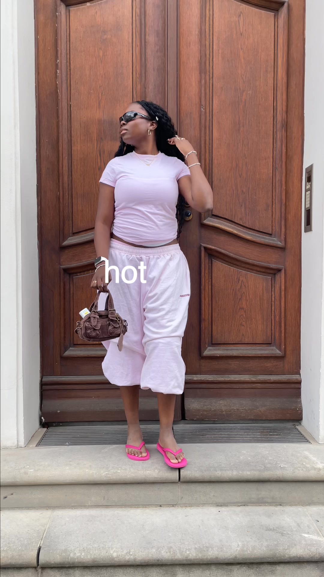 A young Black woman stands on a stone staircase in front of a large wooden door. She is wearing a light pink t-shirt, light pink wide-leg pants, and bright pink flip-flops. She has on dark sunglasses and is holding a small brown handbag. The word "hot" is overlaid in large white text across the image. The overall aesthetic suggests a casual, trendy, and confident look.