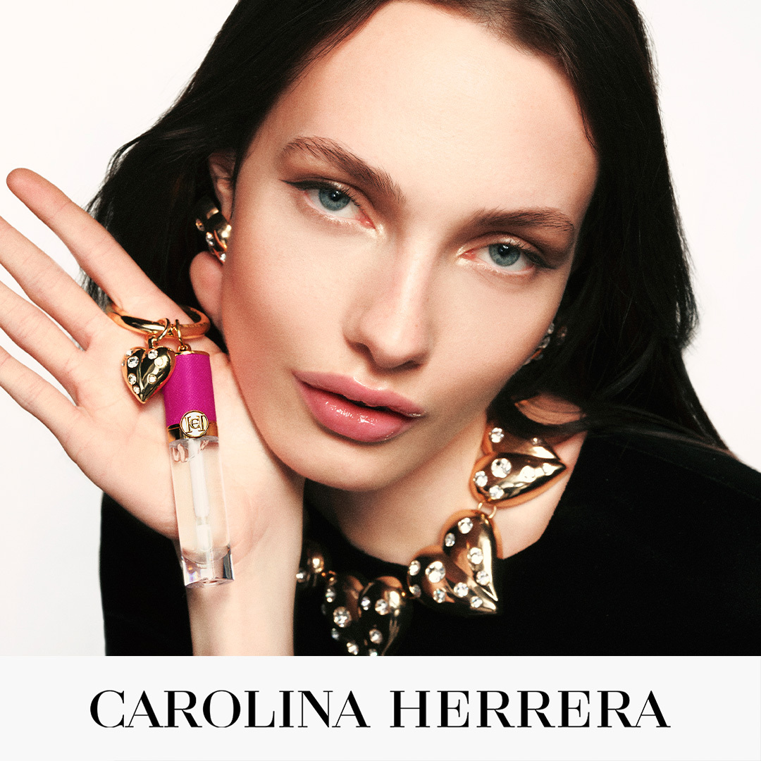 A close-up studio shot features a model with dark hair and striking blue eyes, wearing a black top and a large, ornate gold necklace adorned with clear crystals. She is holding a small, clear lip gloss tube with a vibrant pink cap, which is attached to a gold bracelet with a heart-shaped charm. The text "CAROLINA HERRERA" is displayed prominently in large, white, sans-serif font at the bottom of the image against a white background. The hook, "The obsession is real.", is positioned above the brand name, inviting intrigue.
