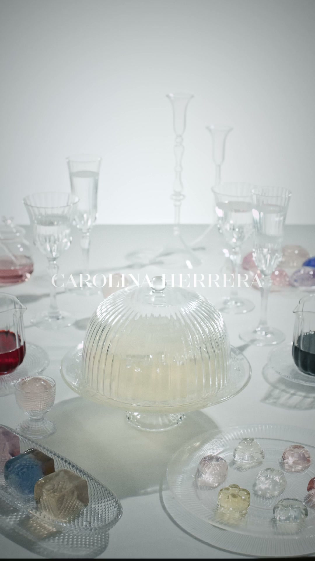 A close-up, high-angle shot of a table setting with various glassware and decorative elements. In the foreground, a clear glass cake stand holds a pale yellow dessert under a ribbed glass dome. To the left, a small glass pitcher contains a dark red liquid, and to the right, a clear glass plate displays several colorful, jewel-like candies. The Carolina Herrera logo is subtly visible in the background, overlaid on the scene.
