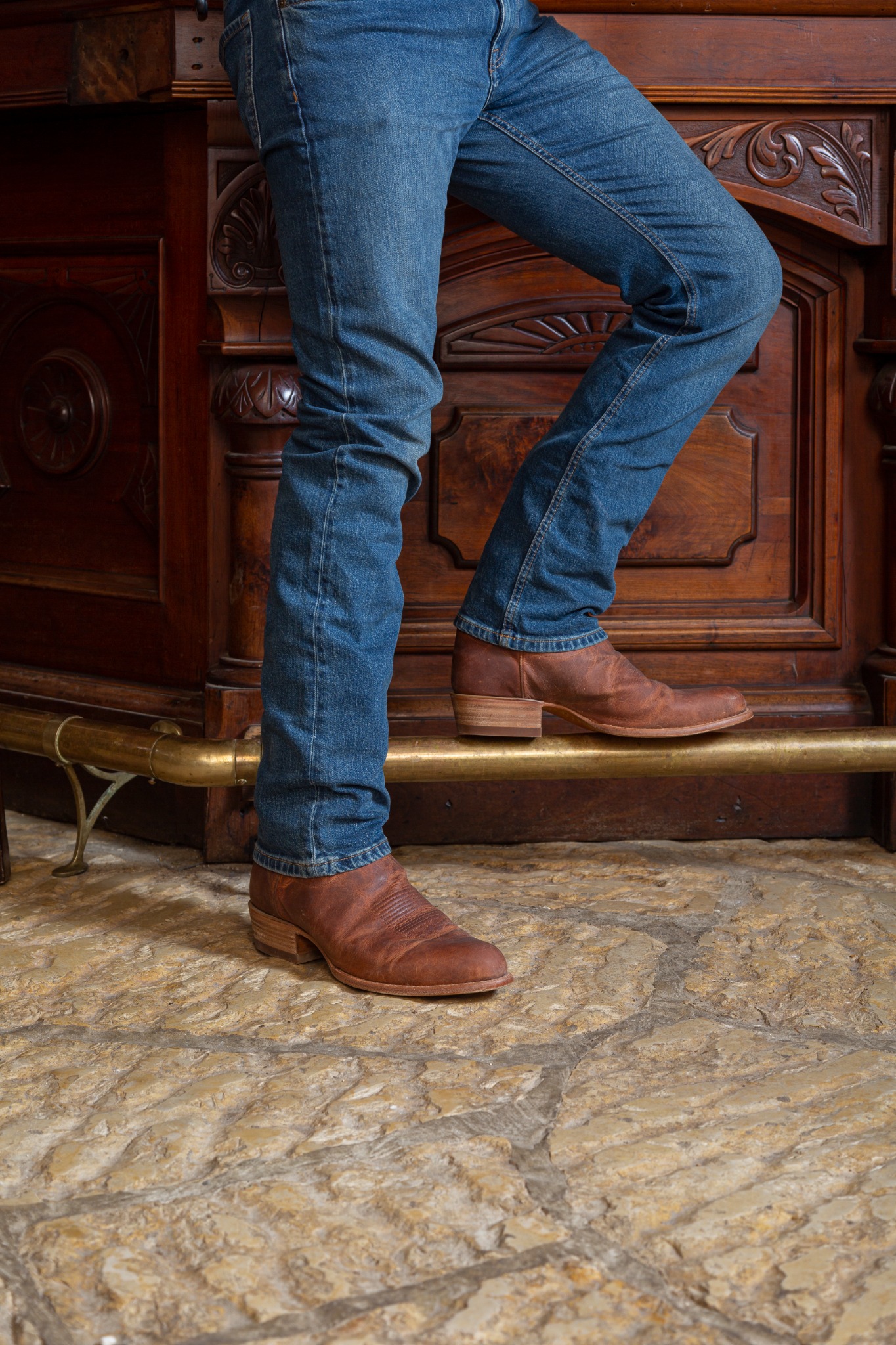 A close-up shot focuses on the lower legs and feet of a person standing in front of a dark, ornate wooden structure, possibly a bar or a piece of furniture. The person is wearing blue denim jeans and brown leather cowboy boots. The boots have a distinctive stitching pattern on the vamp and a stacked heel. The floor is made of rough, uneven stone tiles. Text overlayed on the image reads "The Cartwright in Scotch."