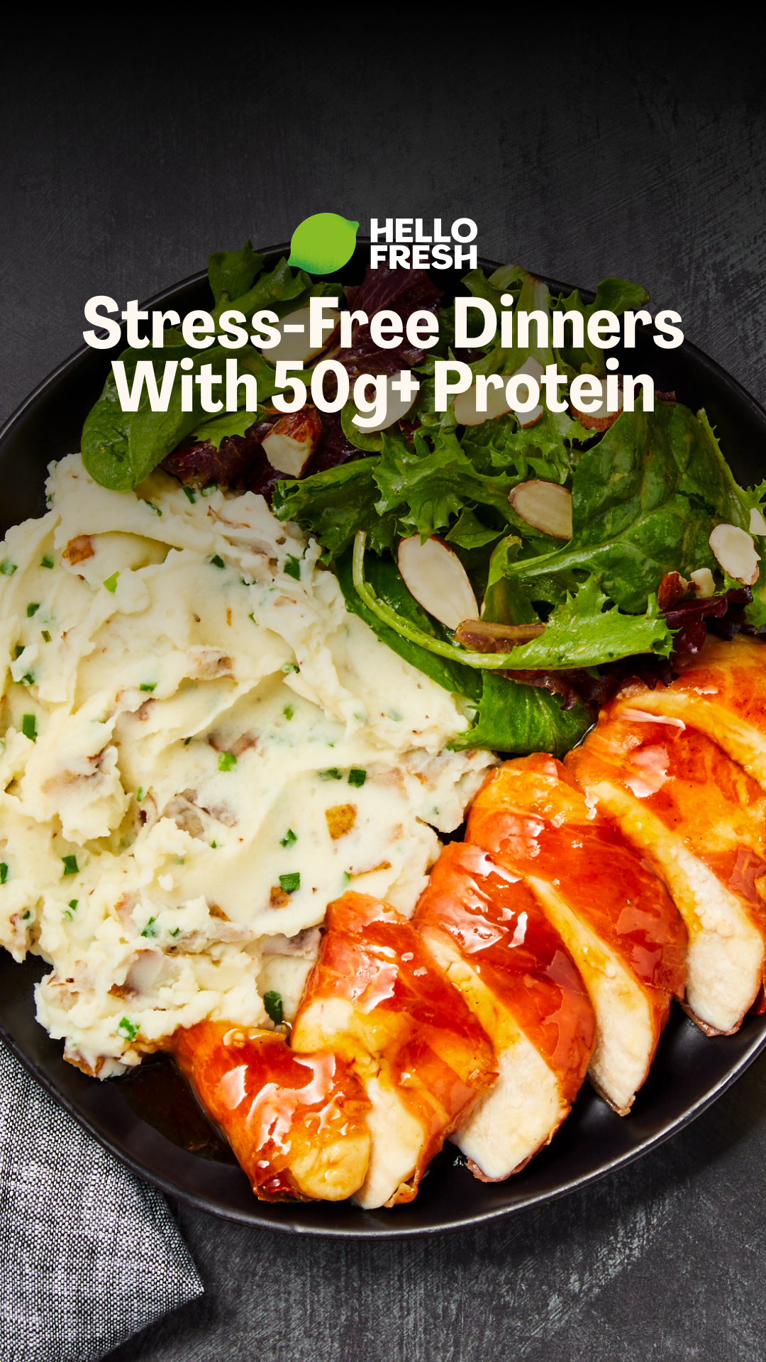 A close-up, overhead shot shows a black plate filled with a HelloFresh meal. The meal consists of a generous portion of creamy mashed potatoes with visible chives and potato chunks, a fresh green salad with sliced almonds, and sliced, glazed chicken breast. The HelloFresh logo and the headline "Stress-Free Dinners With 50g+ Protein" are overlaid in white text on the image. The overall presentation is appetizing and highlights the protein content and ease of preparation.