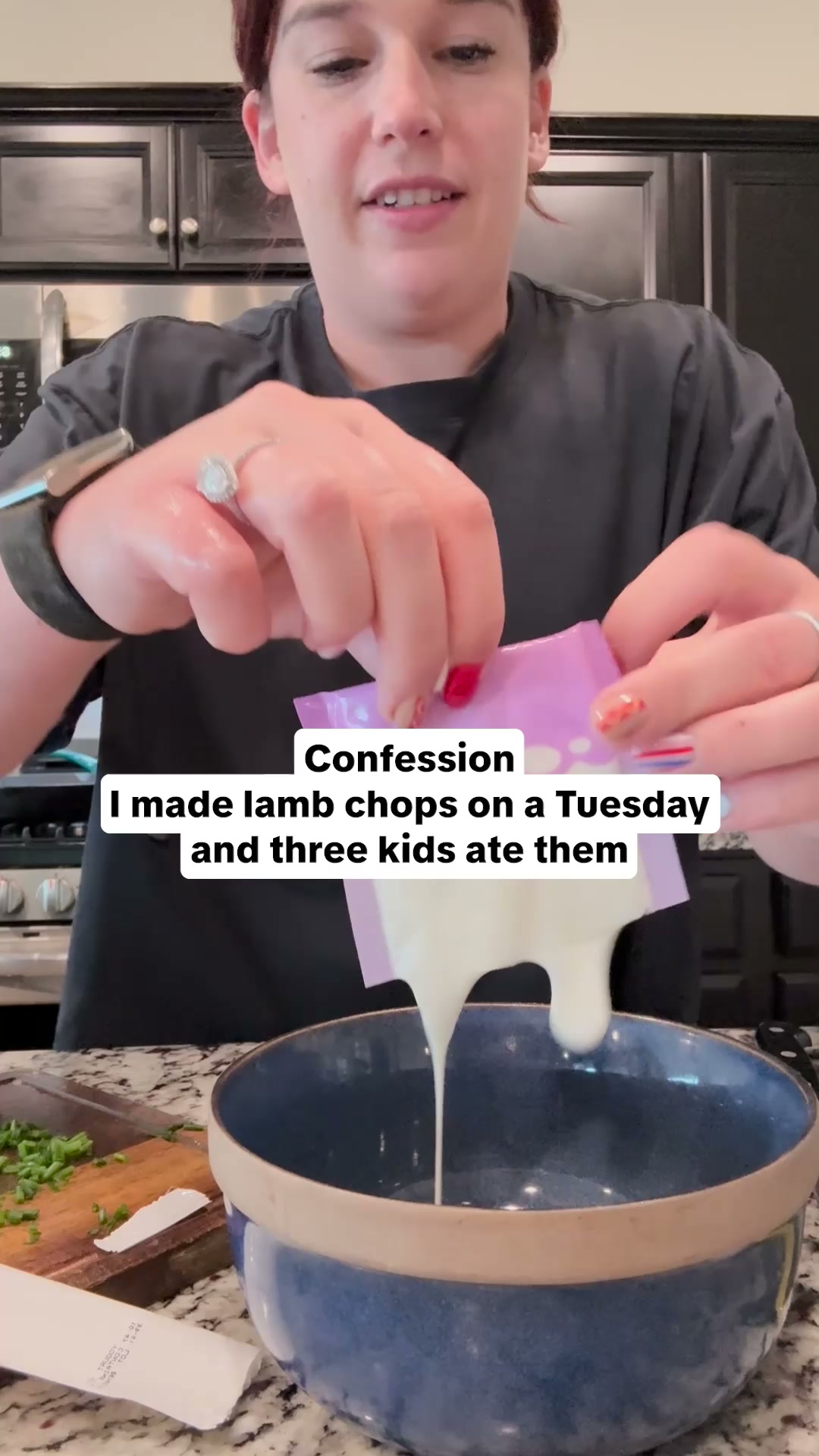 A woman is shown in her kitchen, holding a purple packet and pouring a white liquid into a blue bowl. Text overlays on the image read "Confession" and "I made lamb chops on a Tuesday and three kids ate them." The woman is wearing a dark t-shirt and has rings on her fingers. There is a cutting board with chopped green onions to the left of the bowl.