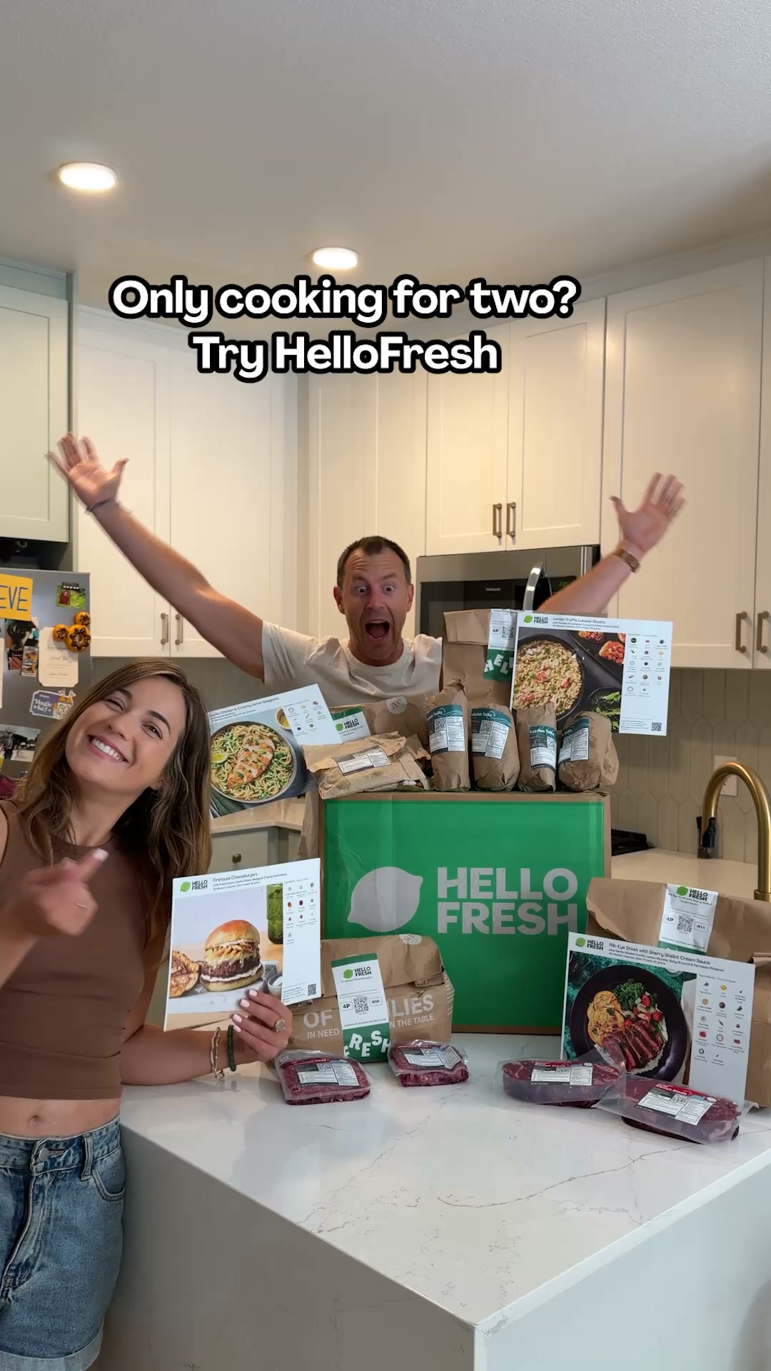 A man and a woman are excitedly posing in a kitchen with a large HelloFresh box and meal ingredients. The woman is pointing towards the camera, and the man has his arms raised in the air. Overlay text on the image asks, "Only cooking for two? Try HelloFresh." The scene showcases the contents of a HelloFresh meal kit, including packaged meats, recipe cards, and bags of ingredients, suggesting convenience and ease of meal preparation for couples.