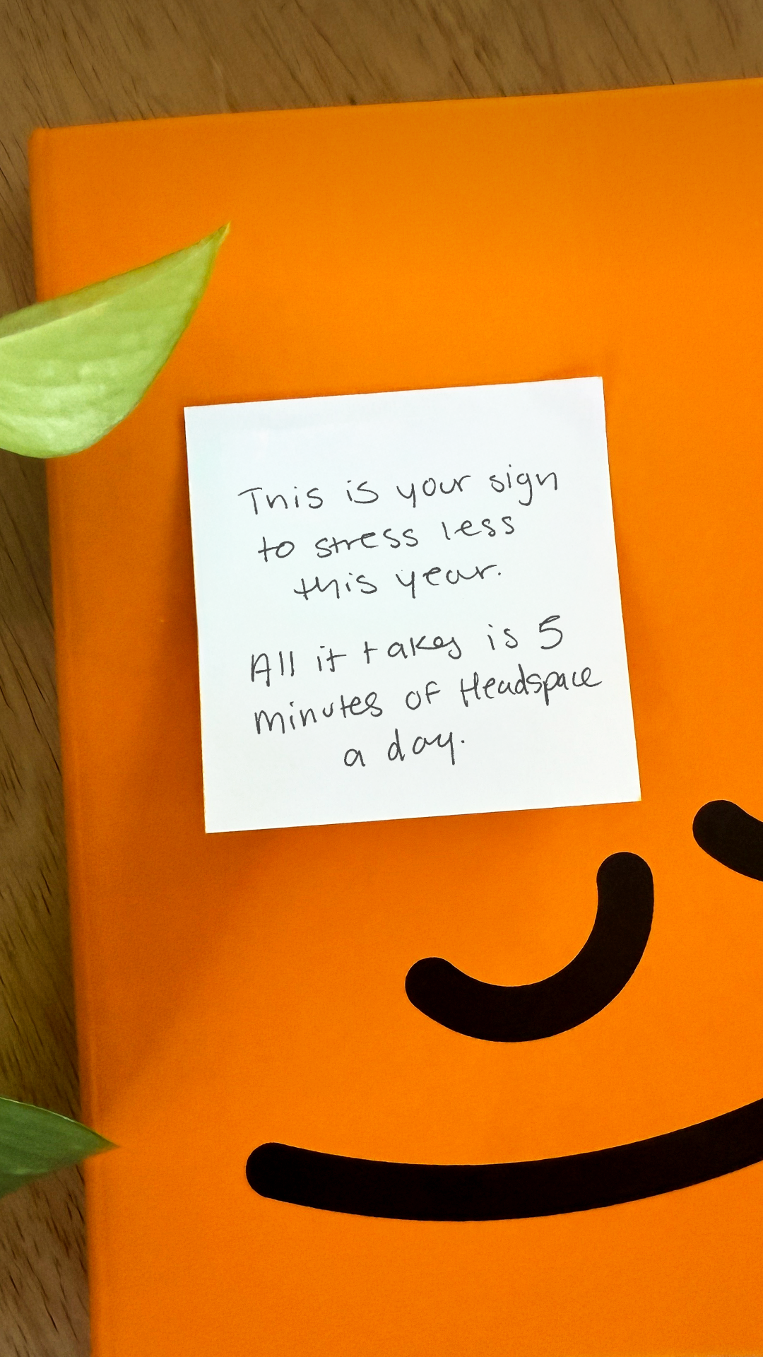 A white sticky note with handwritten text is placed on an orange notebook. The text reads: "This is your sign to stress less this year. All it takes is 5 minutes of Headspace a day." The notebook has a simple black smiley face drawn on it. A green leaf is visible in the top left corner, and another in the bottom left, adding a touch of nature to the scene. The overall aesthetic is clean and minimalist, with the bright orange providing a cheerful backdrop.
