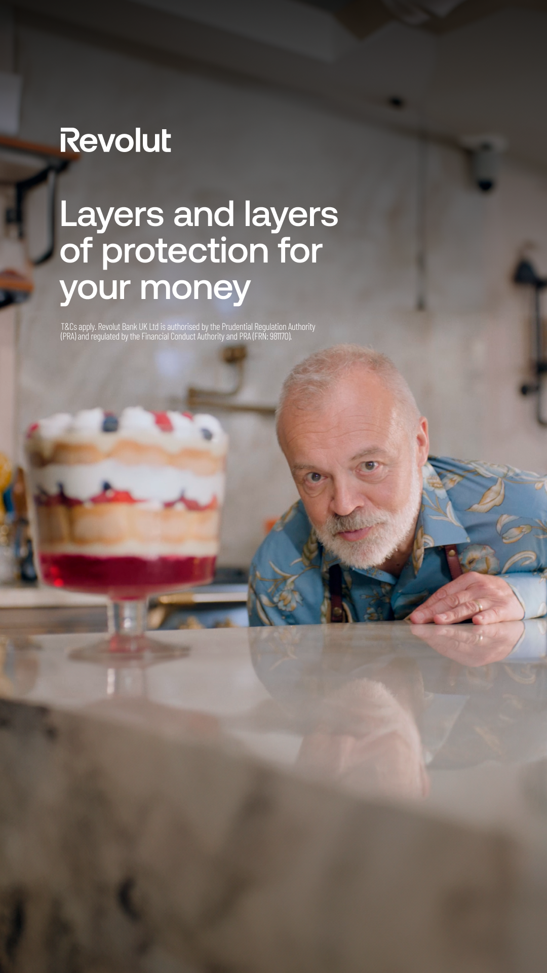 A man with a grey beard and a blue floral shirt is looking at the camera. In front of him is a large glass of trifle. The Revolut logo is in the top left corner, and the headline "Layers and layers of protection for your money" is displayed in white text. Below the headline, there is smaller text with terms and conditions. The overall scene is set in what appears to be a cafe or bar.