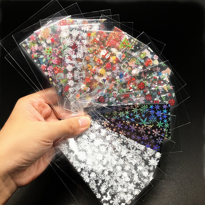 A hand holds up several clear plastic sheets, each containing various Christmas-themed nail art stickers. The stickers feature designs like snowflakes, snowmen, Santa Claus, Christmas trees, bows, and poinsettias. The sheets are fanned out, showcasing a variety of patterns and colors, including some with holographic or glitter effects against dark backgrounds.