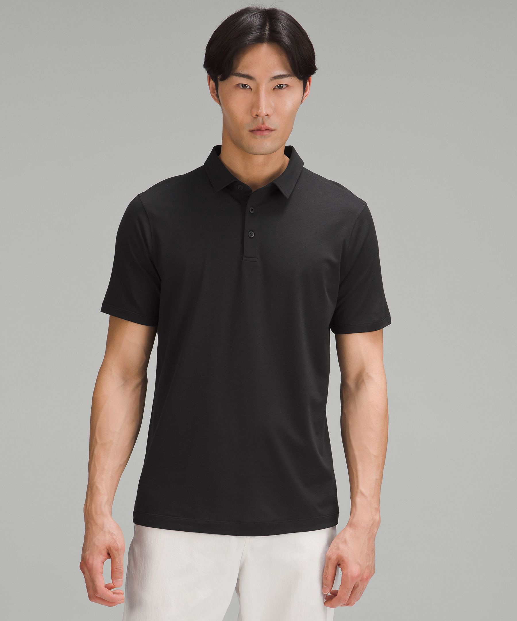 A man of Asian descent stands against a plain grey background, wearing a black Lululemon polo shirt and light-colored shorts. He looks directly at the camera with a neutral expression. The text "Whatever your goals are this season, find the gear that’ll keep you moving." is overlaid on the image. The overall presentation is clean and focuses on the product and its aspirational message of movement and goal achievement.