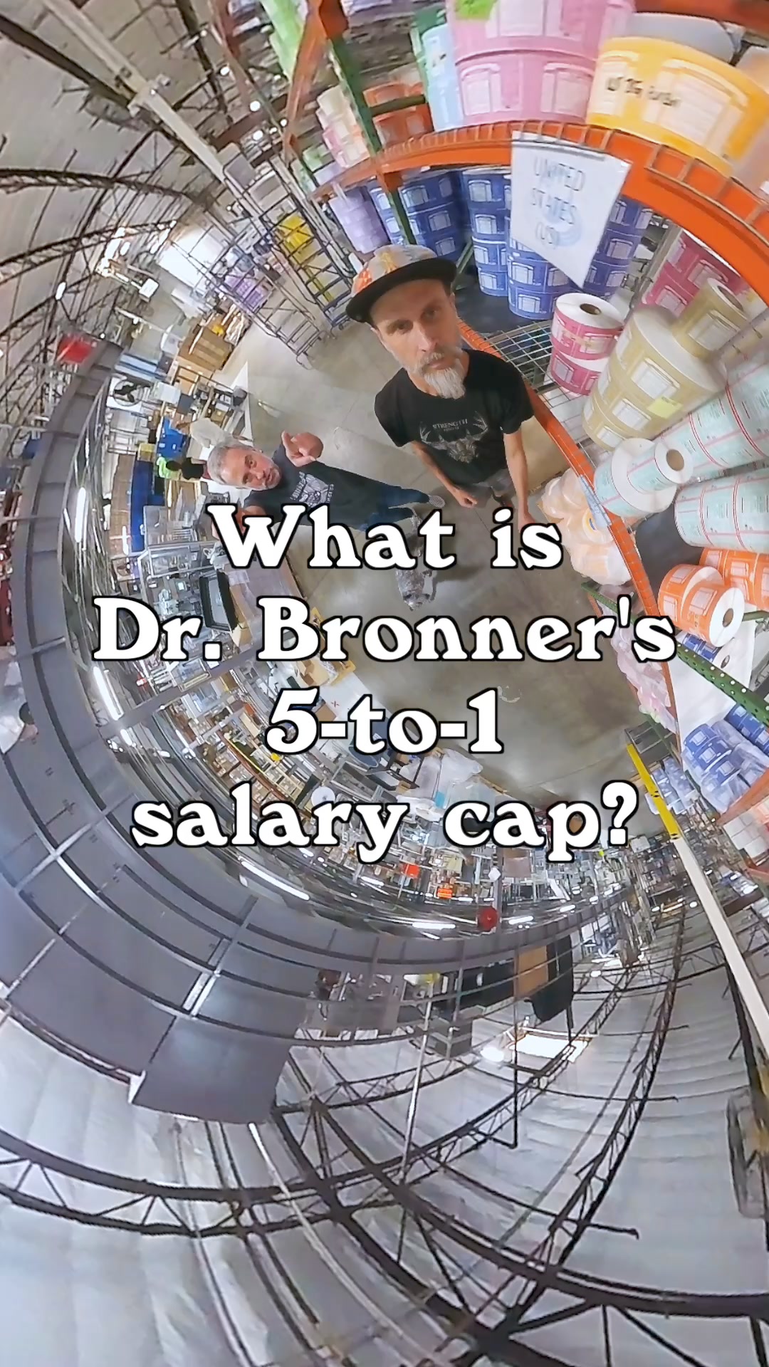 A wide-angle, fisheye lens shot shows the interior of a warehouse with shelves stocked with rolls of labels. Two men are visible in the center of the frame. White text overlaid on the image asks, "What is Dr. Bronner's 5-to-1 salary cap?" The copy "Let's talk about our 5-to-1 salary cap." is also present.