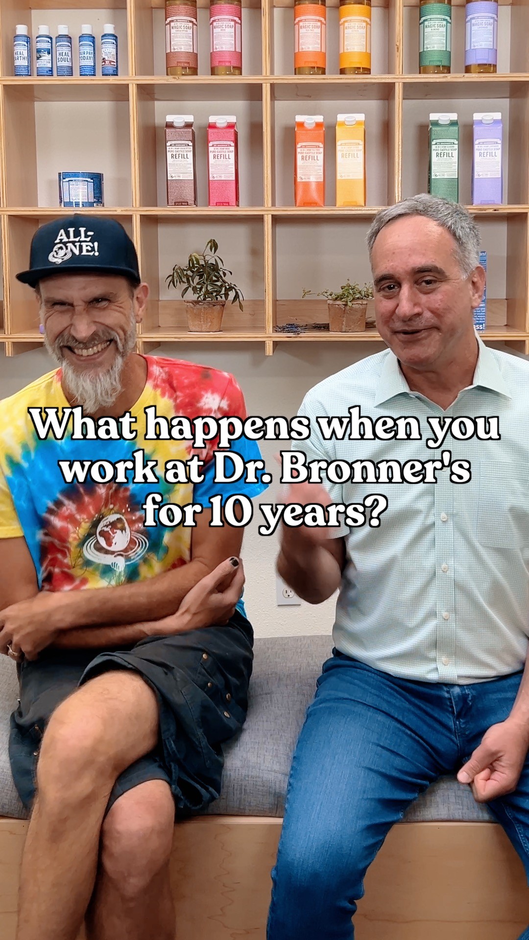 Two men are sitting on a couch in front of shelves stocked with Dr. Bronner's products. The man on the left is wearing a tie-dye t-shirt and a baseball cap, and he is smiling widely with his eyes squinted. The man on the right is wearing a light blue collared shirt and is looking at the camera with a slight smile. White text overlaid on the image asks, "What happens when you work at Dr. Bronner's for 10 years?"