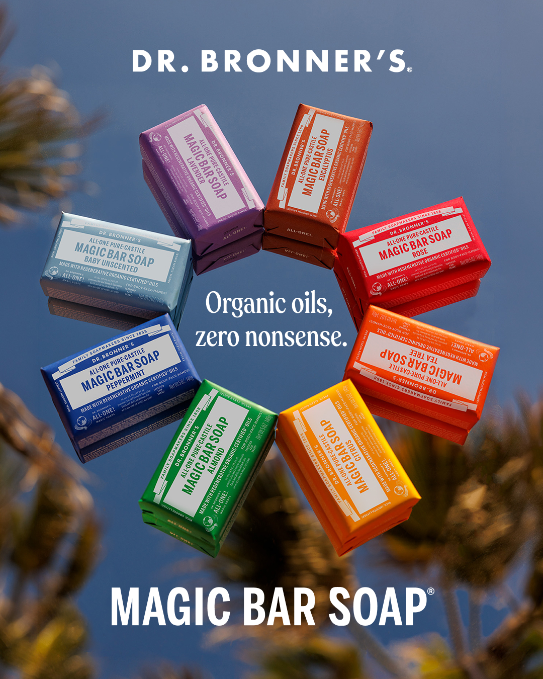 A circular arrangement of seven colorful Dr. Bronner's Magic Bar Soaps is shown against a blurred background of palm fronds and a blue sky reflected on a mirrored surface. The soaps are in various scents like Lavender, Eucalyptus, Rose, Tea Tree, Citrus, Almond, and Unscented. The brand's logo, "DR. BRONNER'S", is displayed prominently at the top, and the tagline "Organic oils, zero nonsense." is overlaid in the center. At the bottom, the product name "MAGIC BAR SOAP" is written in large white letters.