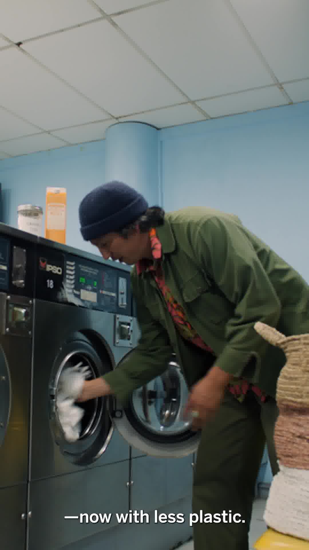 A person wearing a beanie and a green jacket is putting laundry into a washing machine. The scene is set in a laundromat with blue walls and fluorescent lighting. On-screen text at the bottom reads "-now with less plastic."