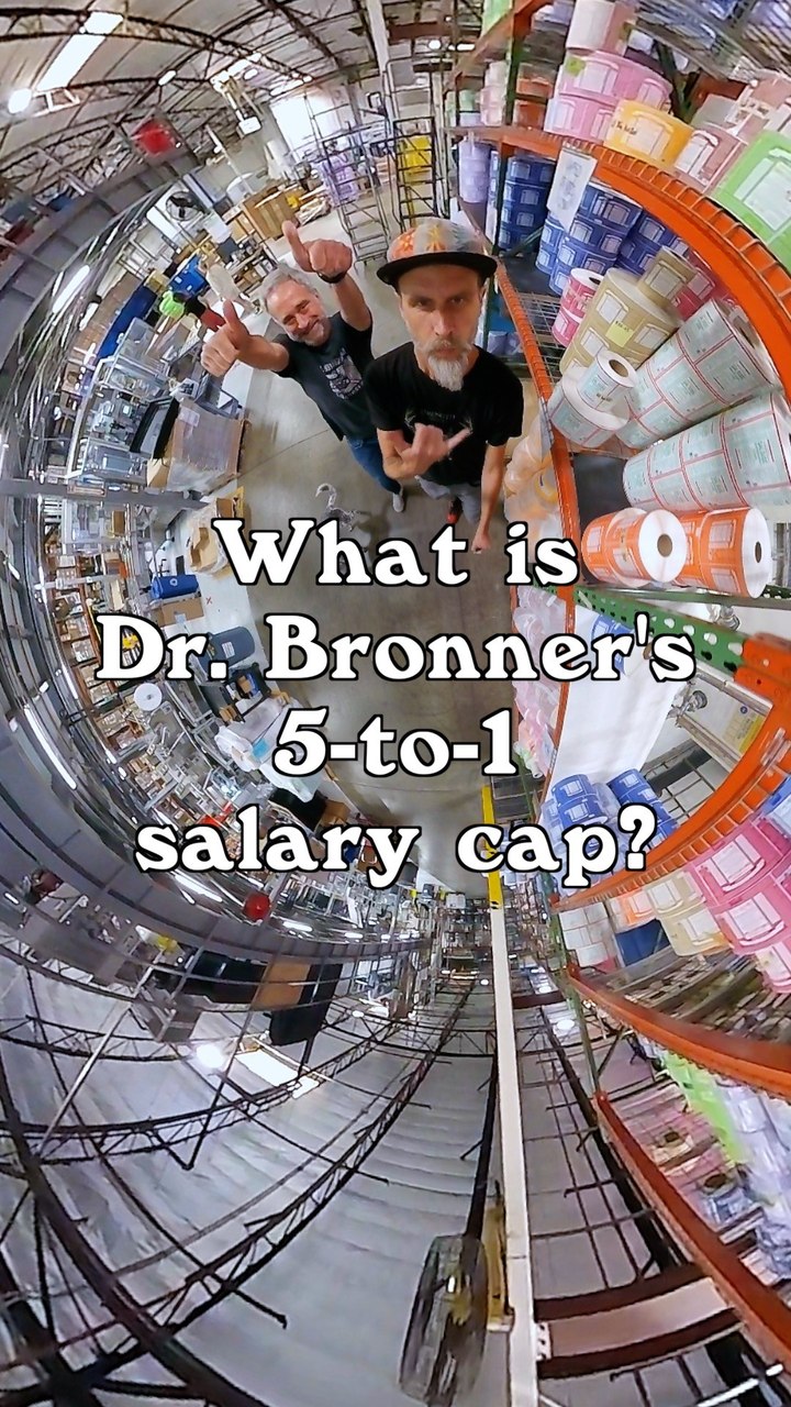 A fisheye lens view of two men standing in a warehouse aisle filled with shelves of products. The man on the left is smiling and giving a thumbs up, while the man on the right looks directly at the camera with a serious expression and makes a hand gesture. Large white text overlays the image, asking "What is Dr. Bronner's 5-to-1 salary cap?" The overall impression is informative and slightly quirky, hinting at a unique business practice.