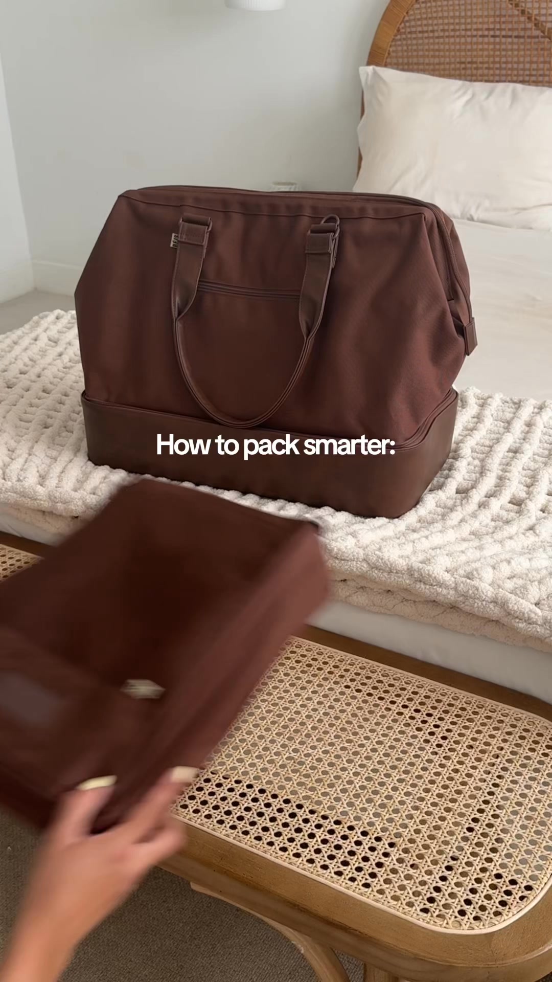 A brown duffel bag is placed on a bed with a textured cream blanket. A hand is reaching into the frame from the left, holding a brown packing cube. White text overlaid on the image reads "How to pack smarter:". The scene is set in a bedroom with a wicker headboard visible in the background.