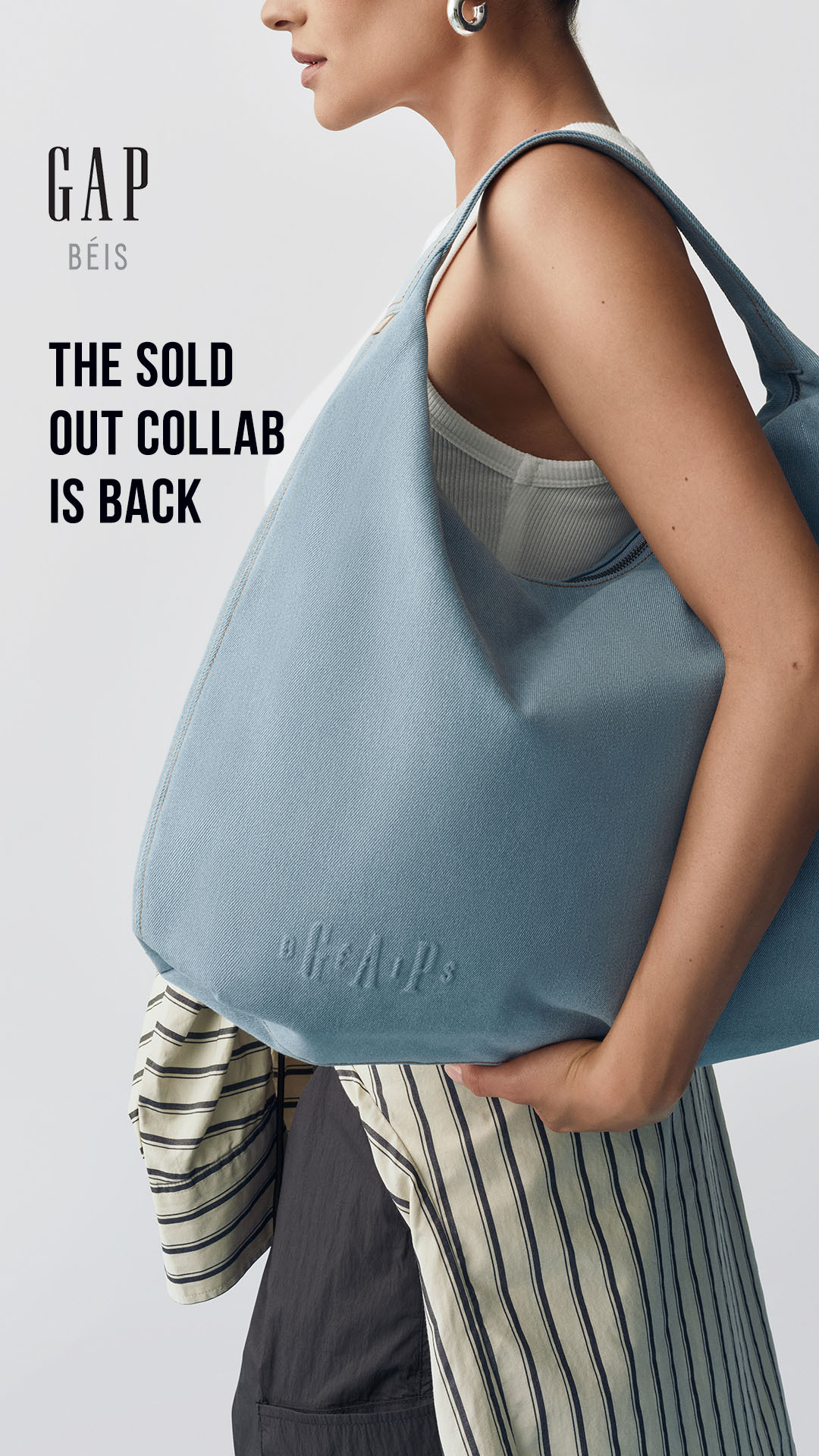 A woman is shown from the chest up, holding a light blue denim tote bag. The bag has the embossed logo "GAP x BÉIS" on it. The text "GAP BÉIS" is displayed in the upper left corner, followed by the headline "THE SOLD OUT COLLAB IS BACK" below it. The woman is wearing a white ribbed tank top and large silver hoop earrings. The background is a plain light gray.
