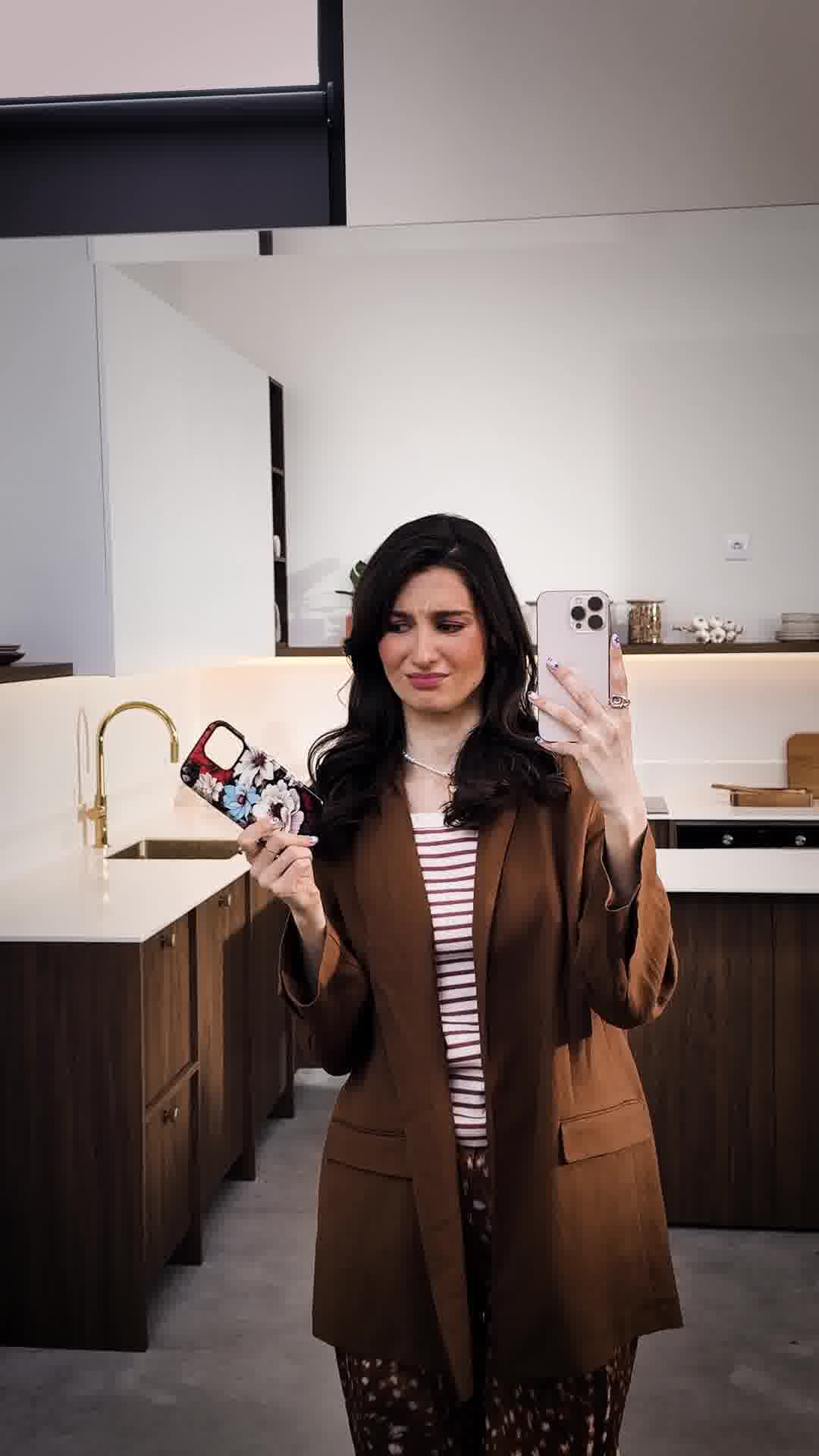 A woman in a brown blazer and striped shirt stands in a modern kitchen, holding up a phone in one hand and a floral phone case in the other. She has a slightly displeased expression. Text overlays the image, listing three reasons why someone might order multiple phone cases from the brand BURGA. The ad promotes a '4 for the price of two' offer and directs viewers to burga.com.