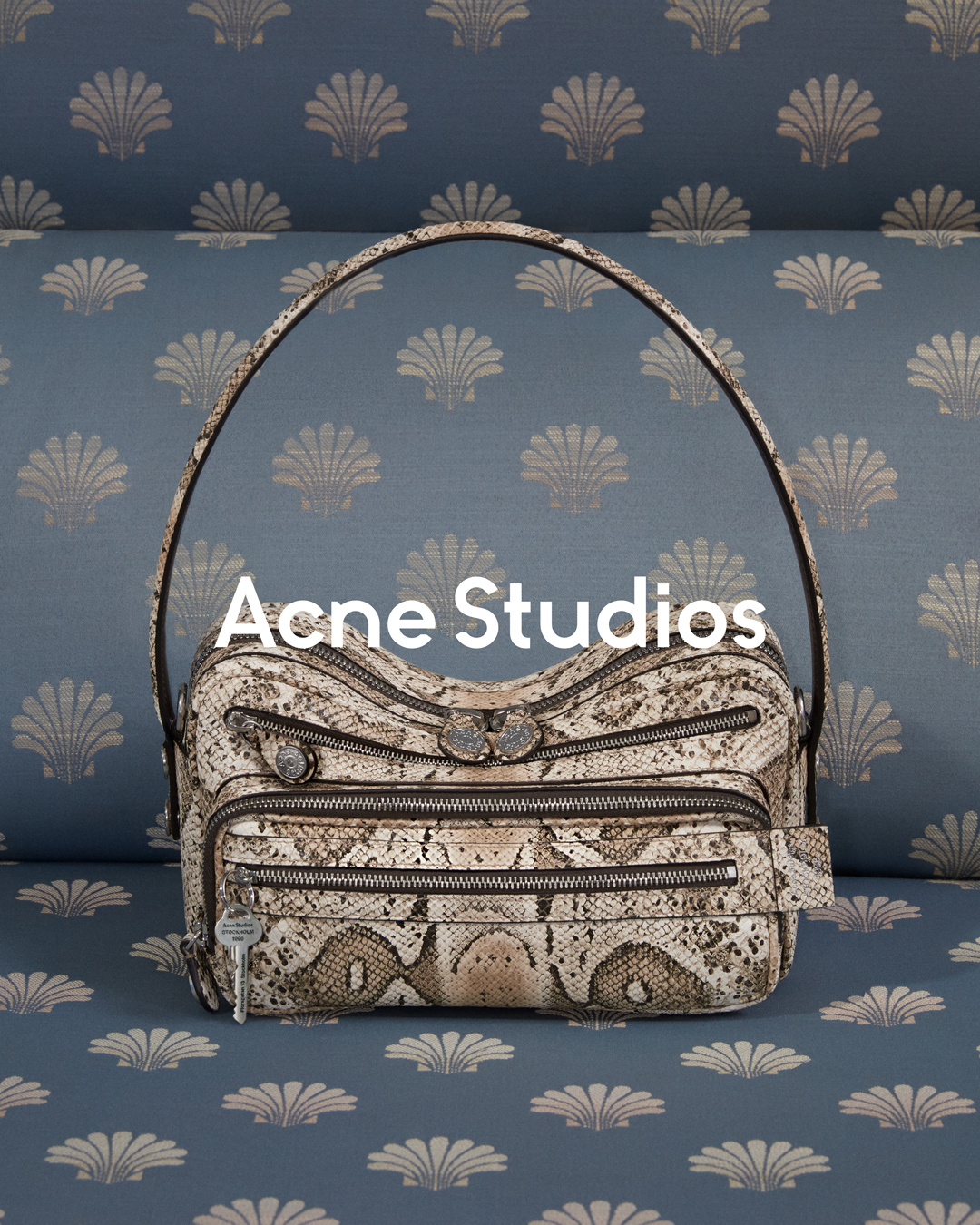 A close-up shot of a textured, multi-zippered handbag with a prominent Acne Studios logo on the front. The bag is placed on a blue upholstered surface with a repeating shell pattern. White text overlaying the image reads "2026年秋冬 カメロバッグ", which translates to "Fall/Winter 2026 Camello Bag". The accompanying Japanese copy describes the Camello bag as a reinterpretation of classic codes with a youthful and free sensibility, available in new seasonal colors.