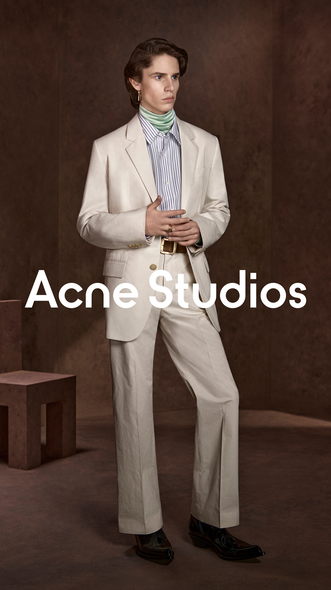 A young man in a cream-colored suit stands against a textured brown background. He is wearing a striped shirt with a light green turtleneck underneath. The Acne Studios logo is prominently displayed in white text across the lower half of the image. The text overlay announces "Fall Winter 2026 Menswear" and provides a brief description of the collection's aesthetic, along with information on availability.