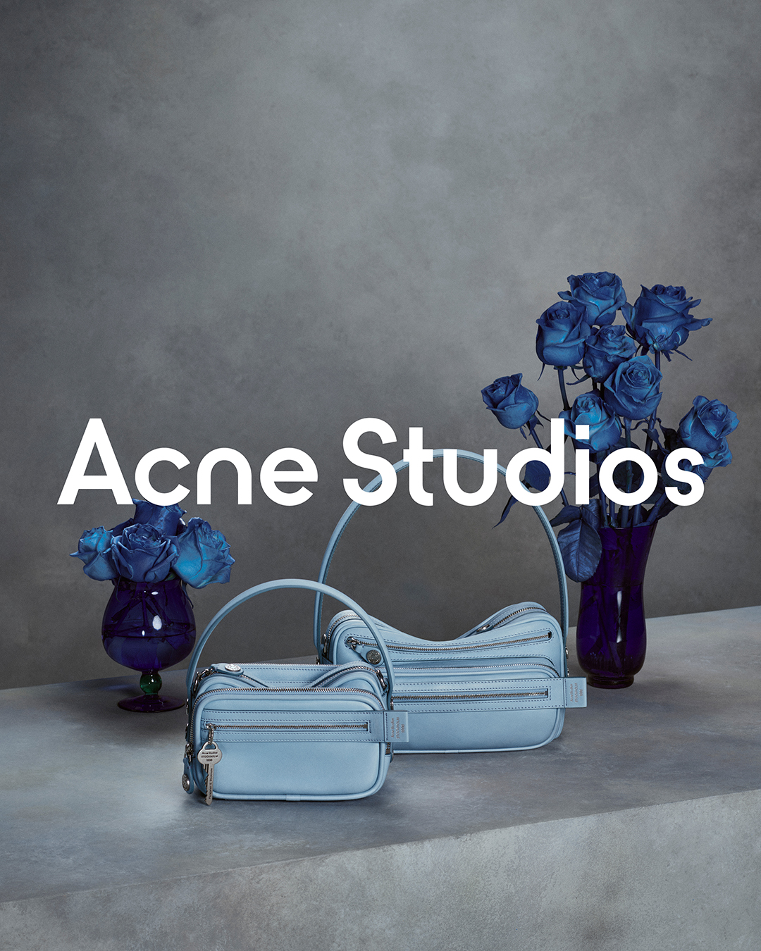 A studio shot features two light blue leather handbags from Acne Studios against a textured grey background. Blue roses in a dark purple vase and a smaller blue vase add a pop of color. The brand name "Acne Studios" is displayed in large white text across the center of the image. The hook, "FW26 Camero bag," is positioned at the top left, directly addressing the viewer.