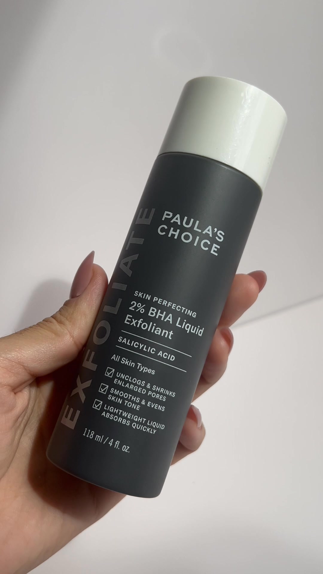 A hand holds a dark grey bottle of Paula's Choice 2% BHA Liquid Exfoliant against a light grey background. The bottle has white text that reads "EXFOLIATE" vertically on the left side and "PAULA'S CHOICE" horizontally near the top. Below that, it lists "SKIN PERFECTING 2% BHA Liquid Exfoliant" and "SALICYLIC ACID". Bullet points highlight benefits like "UNCLOGS & SHRINKS ENLARGED PORES", "SMOOTHS & EVENS SKIN TONE", and "LIGHTWEIGHT LIQUID ABSORBS QUICKLY". The bottom of the bottle shows "118 ml / 4 fl. oz.".