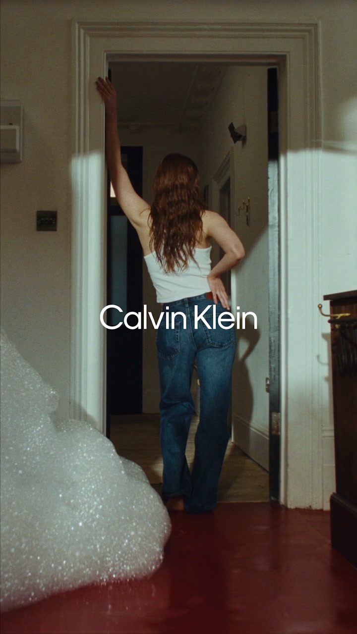 A young woman with long, wavy red hair, wearing a white tank top and blue jeans, stands with her back to the camera, reaching her left arm up towards a doorway. The Calvin Klein logo is prominently displayed in white text on the right side of the image, overlaying the scene. In the foreground, a large pile of white foam bubbles is visible on the left, and the floor is a deep red color. The overall aesthetic is intimate and emphasizes the fit and feel of the jeans.