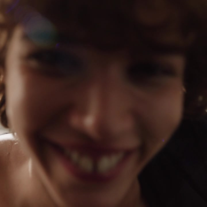 A close-up, slightly blurred shot of a person's face, with a wide, toothy smile. The lighting is soft and warm, highlighting the contours of their face and the texture of their hair. Overlaid on the image is text that reads "Sensualità in pizzo." which translates to "Lace sensuality." The overall impression is intimate and alluring, focusing on the feeling evoked by the product rather than the product itself.