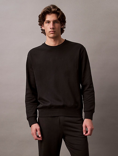 A male model with medium-length brown hair is shown from the chest up, wearing a black crewneck sweatshirt with subtle embossed branding on the chest. He is looking directly at the camera with a neutral expression. The background is a plain, muted grey. The text "Discover new classics from Calvin Klein." is overlaid in white sans-serif font at the top of the image.