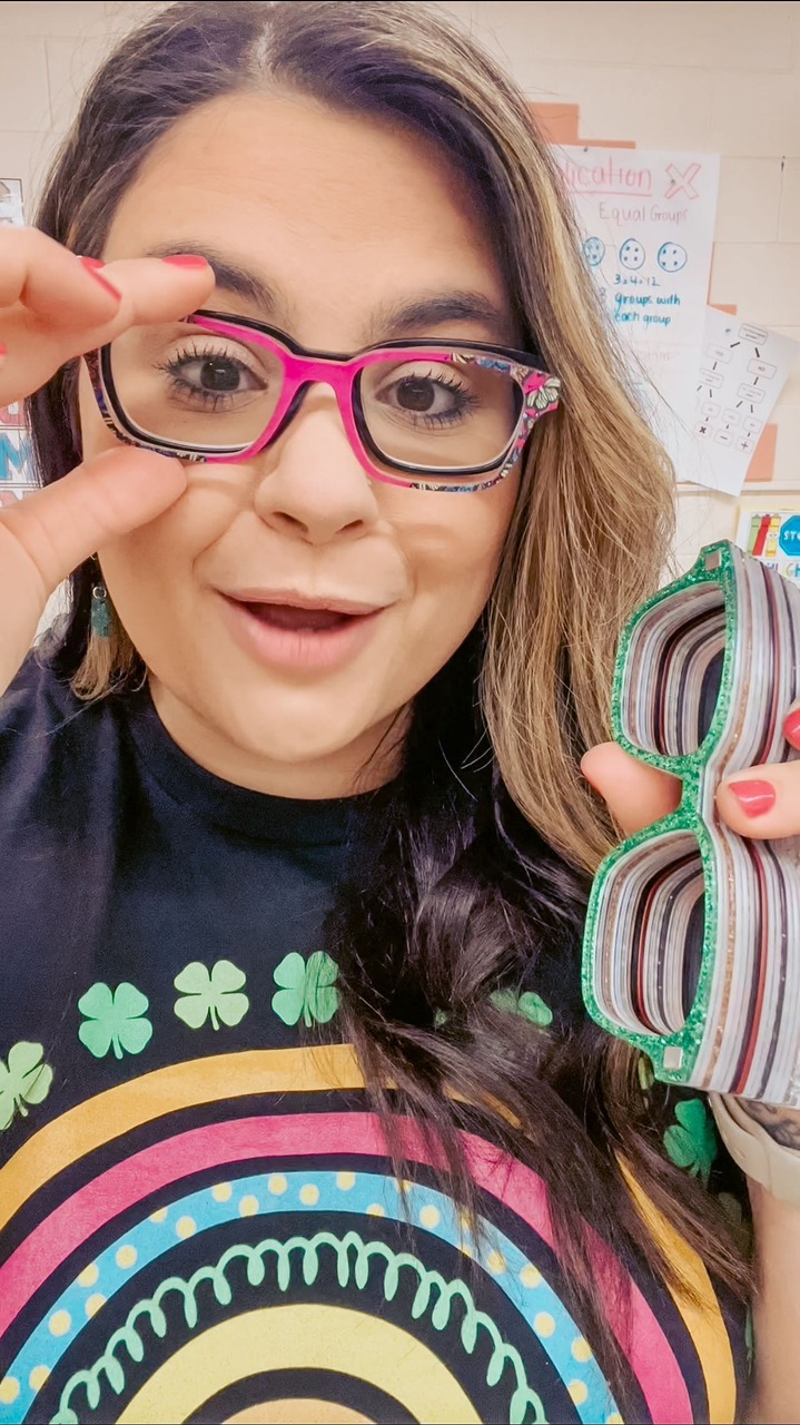 A woman with glasses is holding up a pair of glasses to the camera. She is wearing a black t-shirt with a rainbow and shamrocks on it. The background appears to be a classroom with educational posters. The text overlay says "You guys know I love being extra!" and promotes a discount code for Pair Eyewear. The woman is smiling and looking at the camera.