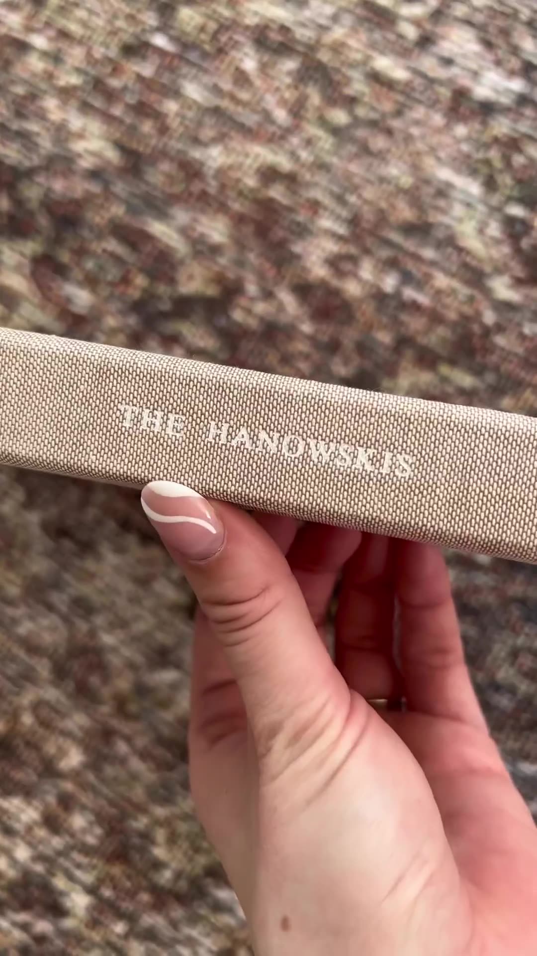 A hand with manicured nails is holding a textured, light-colored book. Embossed white text on the book reads "THE HANOWSKIS." The background is a blurred, mottled surface in shades of brown and beige. The creative uses a contrast hook to suggest the dual functionality of the item as both a wedding guest book and home decor.