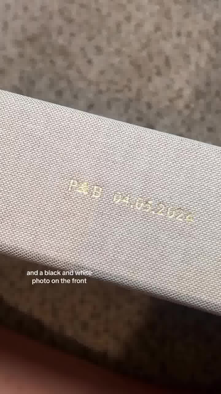 The creative shows a close-up of a textured, light-colored book cover. Embossed gold lettering on the cover reads "P&D 04.05.2024". Below this, white text overlays the image, stating "and a black and white photo on the front". The hook reads, "could not be happier with how this turned out 🥹 wedding photo book from Artifact Uprising!" The overall impression is of a personalized wedding photo album.