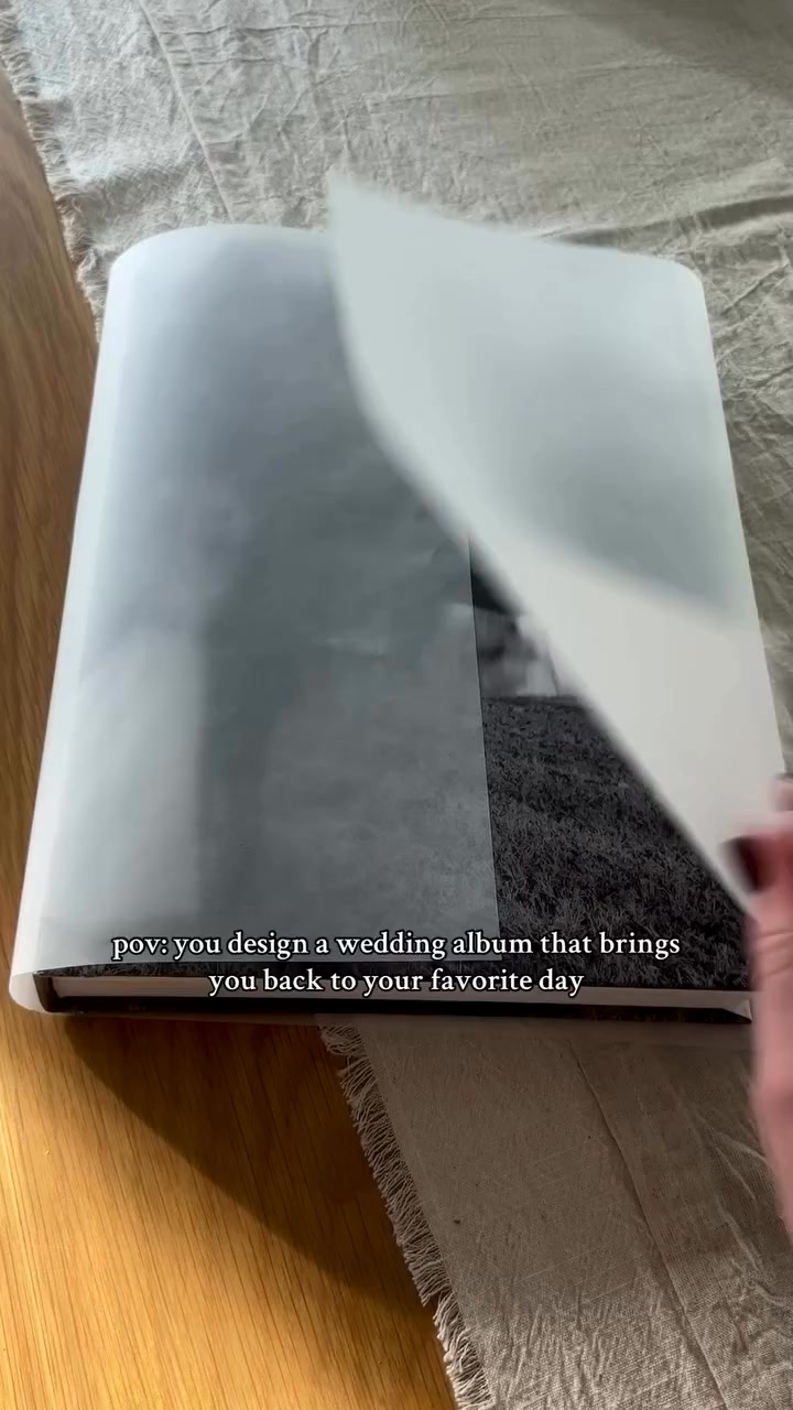A wedding album is shown covered in protective tissue paper, with a hand peeling back a corner. The album is placed on a textured neutral fabric, with a wooden surface visible on the left. Overlay text reads "pov: you design a wedding album that brings you back to your favorite day." The creative focuses on the personal experience of creating a cherished memento.