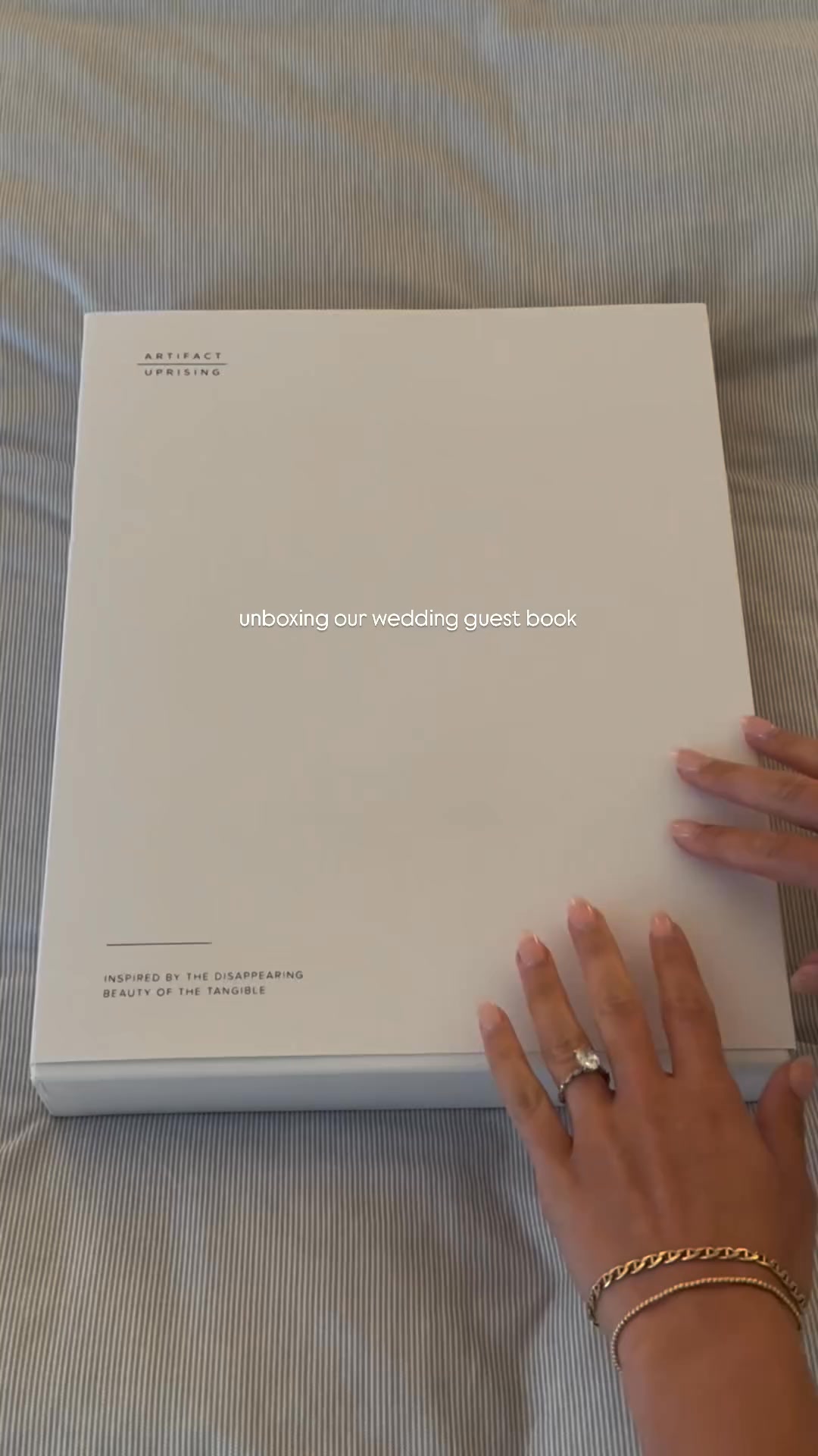 A white box with the Artifact Uprising logo and tagline is shown on a striped grey background. Text overlays the image, reading "unboxing our wedding guest book." The bottom of the box has text that reads "INSPIRED BY THE DISAPPEARING BEAUTY OF THE TANGIBLE." A person's hands are visible on the right side of the frame, touching the box. The creative is a video showing the unboxing of a wedding guest book.