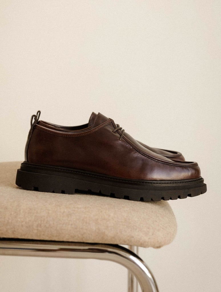 A pair of dark brown leather derby shoes with a thick, lugged sole is displayed on a neutral-colored stool. The shoes are shown from a side profile, highlighting their sleek design and robust construction. The background is a plain, light-colored wall, creating a minimalist aesthetic. The on-screen text emphasizes the contrast between sculpted boots, sleek derbies, and assertive loafers, suggesting a collection that plays with strong lines and rich leather textures for durability and timeless style.