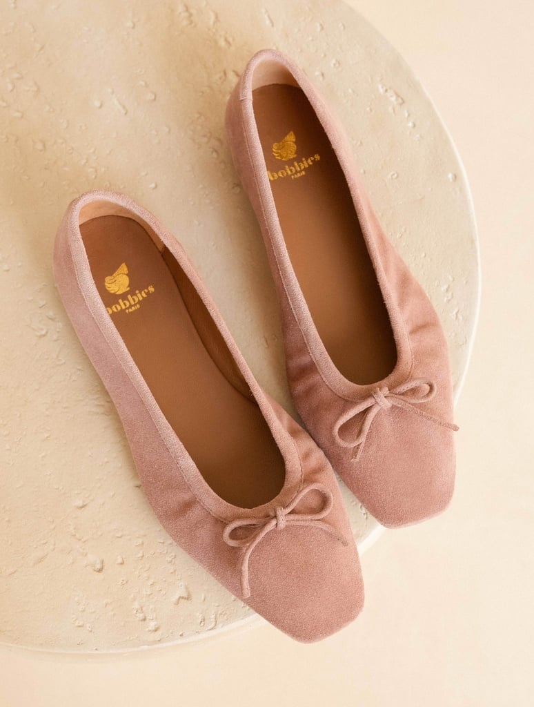 A pair of light pink suede ballet flats are displayed from a top-down perspective against a neutral, textured background. The shoes feature a delicate bow on the toe and the brand's logo, 'bobbies paris', is visible on the insole of each shoe. The on-screen text introduces the new collection with evocative language about the changing season and elegant styles.