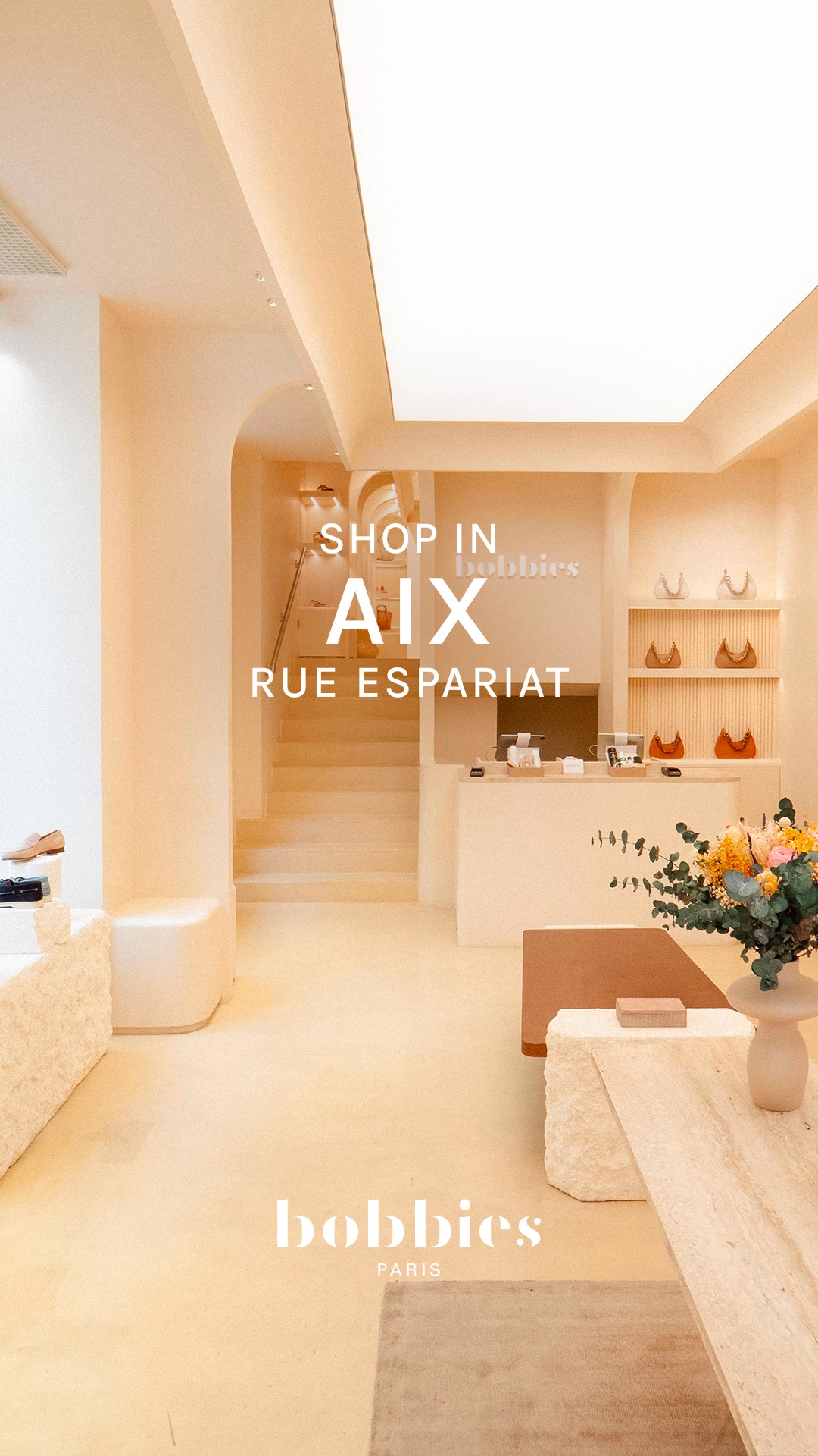 The image shows the interior of a bright, modern boutique with soft, neutral-toned walls and flooring. A staircase is visible in the background, leading to an upper level. The text "SHOP IN AIX" is prominently displayed in large white font, with "bobbies" and "RUE ESPARIAT" also visible. The overall aesthetic is clean and minimalist, highlighting the retail space.