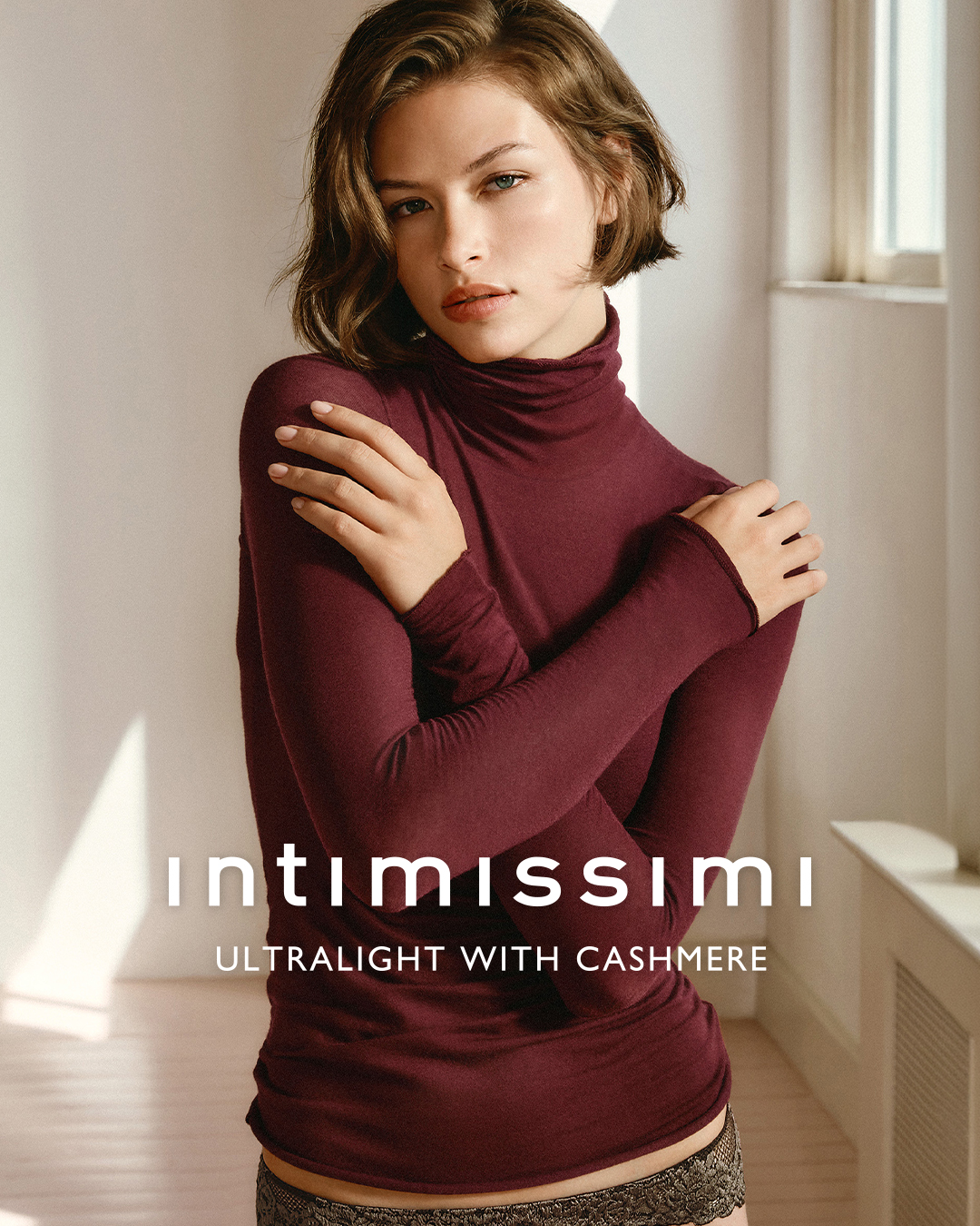 A young woman with short, wavy brown hair and light blue eyes is shown from the chest up, wearing a burgundy turtleneck top and lace-trimmed underwear. She has her arms crossed over her chest, hugging herself. The background is a softly lit room with a window on the right side. White text overlaid on the image reads "intimissimi" in a large, stylized font, followed by "ULTRALIGHT WITH CASHMERE" in a smaller font below. The overall impression is one of comfort and elegance.