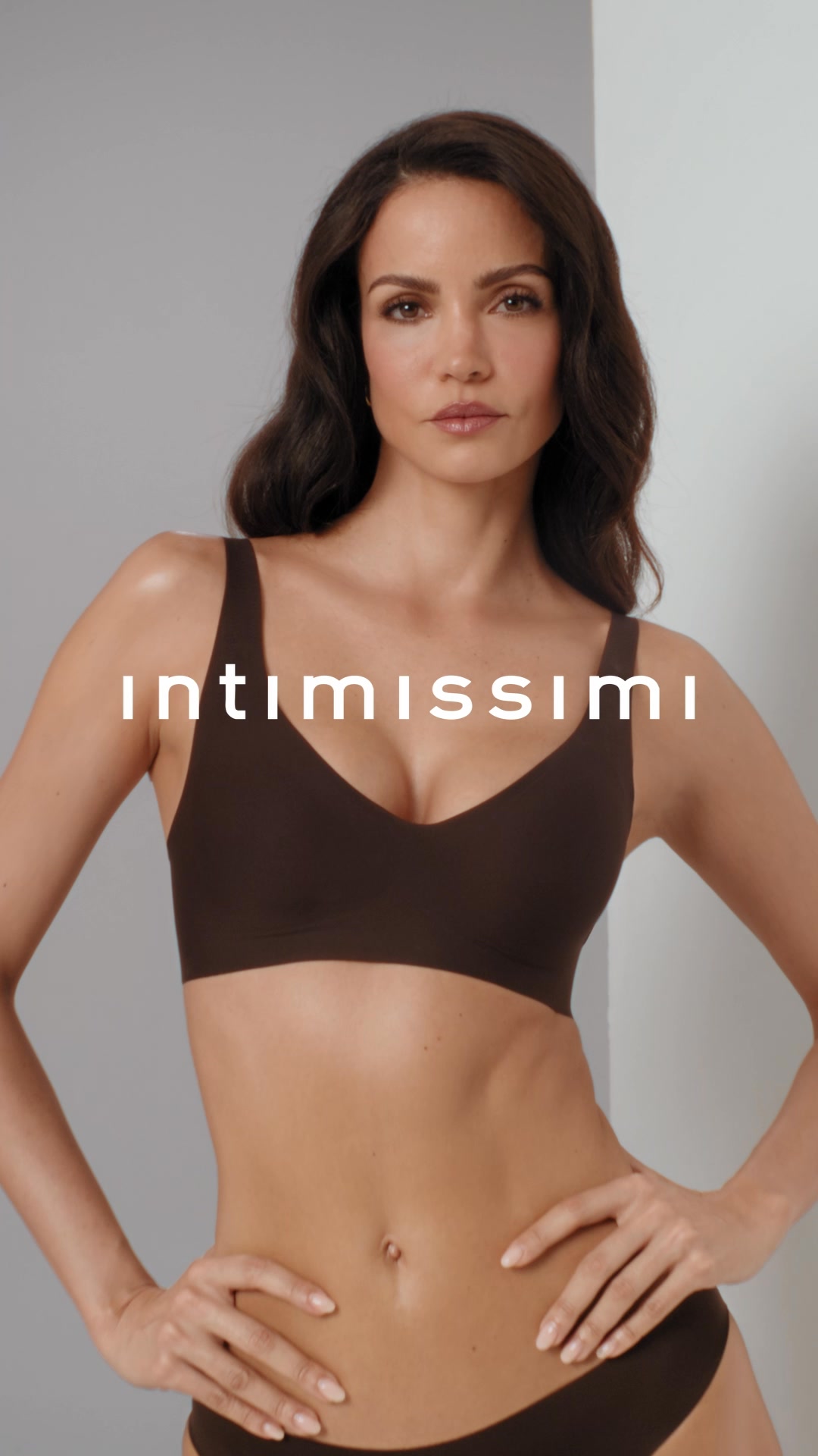 A woman with dark hair is shown from the chest up, wearing a brown bra and matching underwear. She is looking directly at the camera with a neutral expression. White text overlaid on the image reads "intimissimi". The ad highlights the "Aria" bra, emphasizing its clean lines and seamless design for uncompromising comfort, exclusively available at Intimissimi.